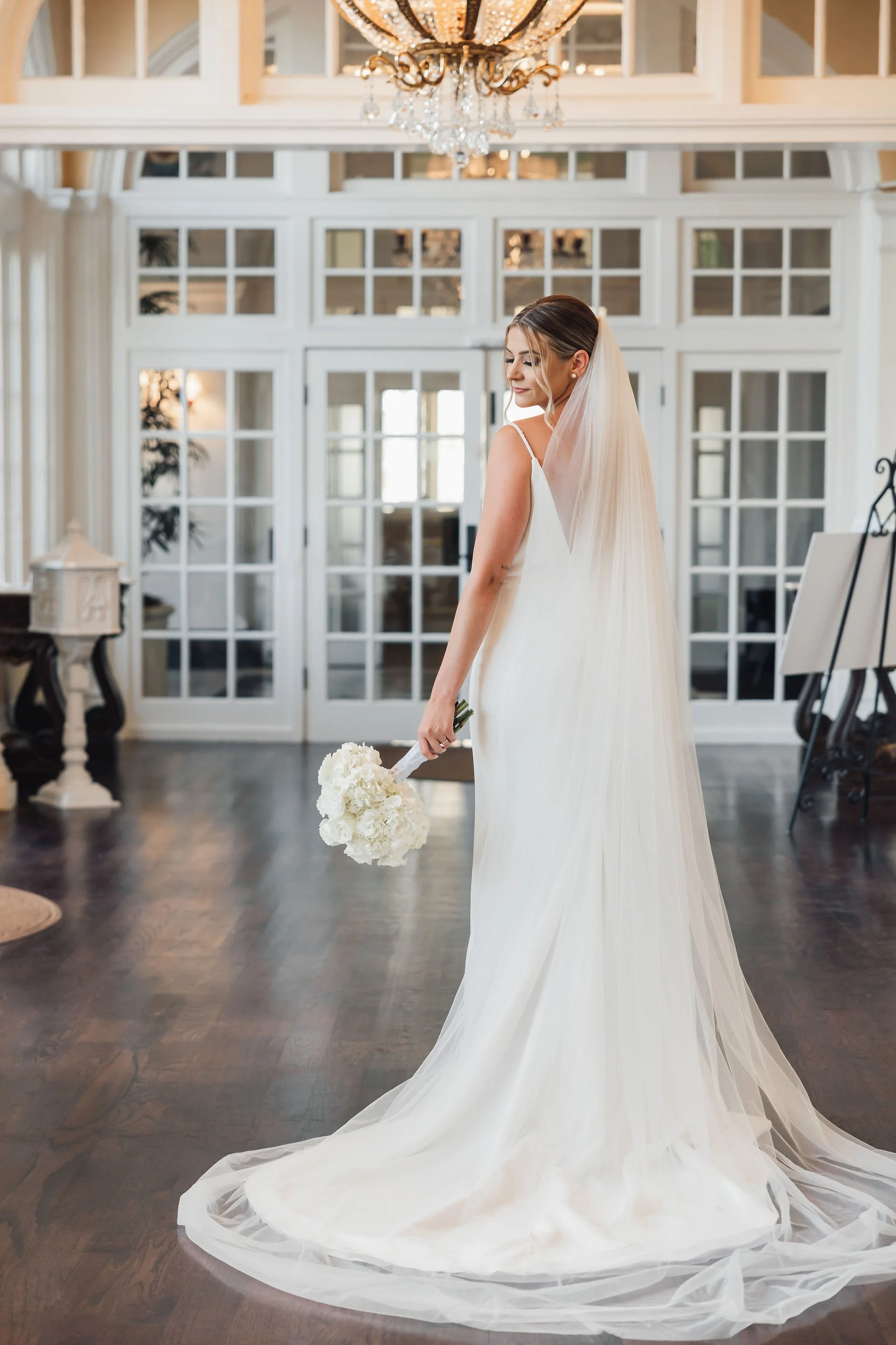 A bride in a white wedding dress with a long train and veil, holding a bouquet of white flowers, standing inside a bright, elegant room with large glass-paneled doors, a chandelier, and dark wooden flooring.