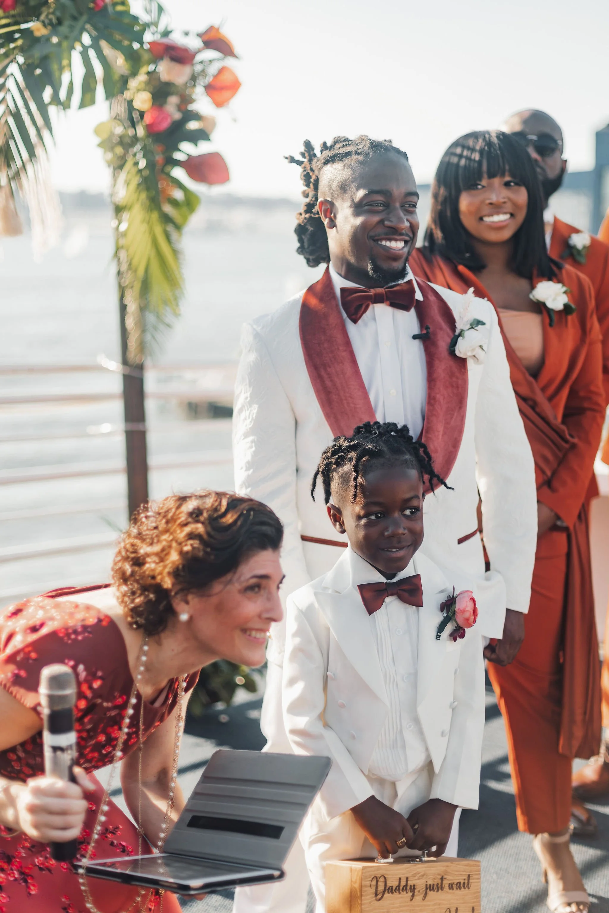 A group of people at a wedding ceremony outdoors near water. The groom is smiling and wearing a white suit with a burgundy bow tie and sash. A young boy dressed in white with a matching bow tie is holding a wooden box, and a woman is speaking into a 