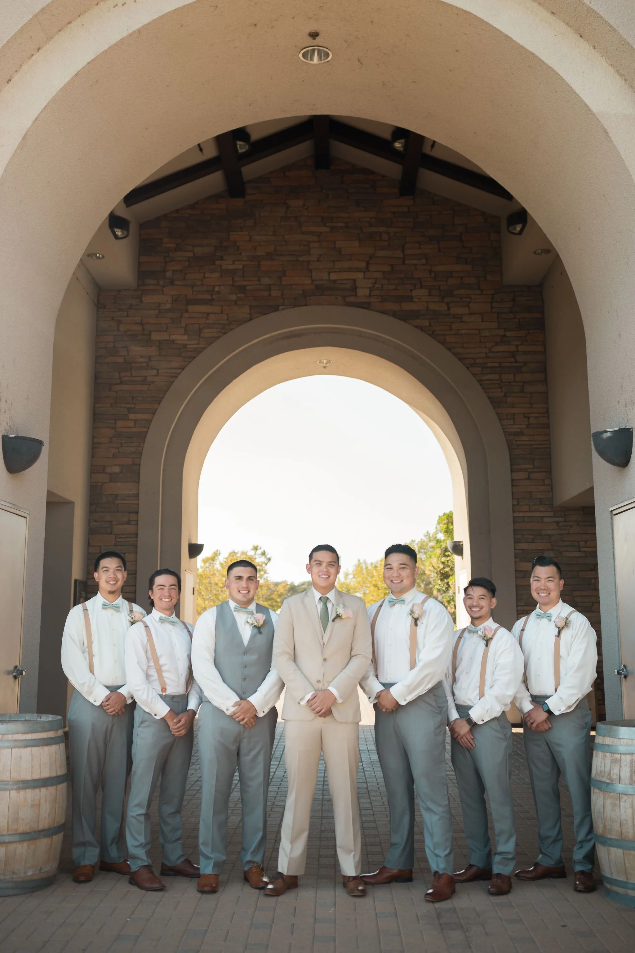 Group of seven men in formal attire standing under an archway, smiling, at a wedding venue.