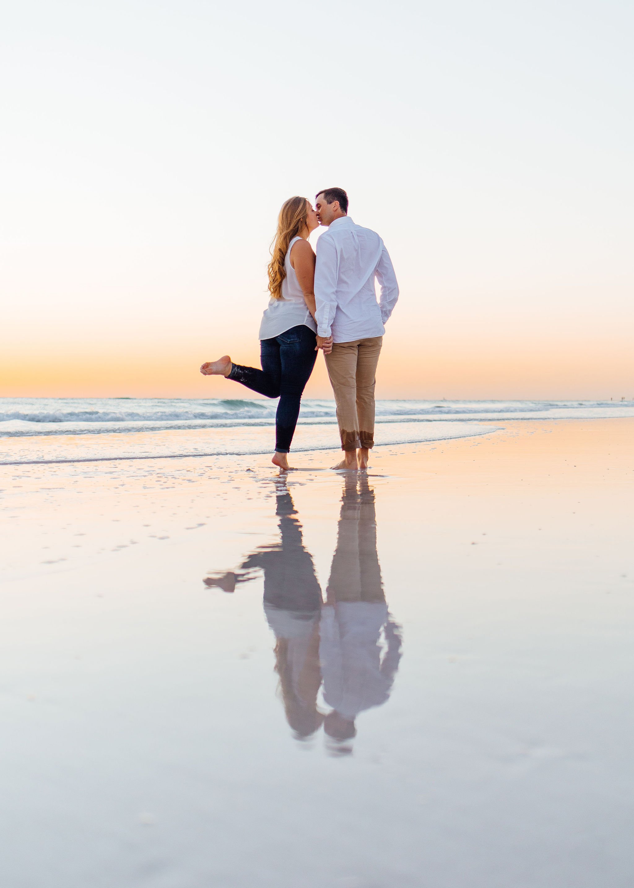A couple holding hands on the beach at sunset, standing in shallow water, with reflections of their figures on the wet sand.