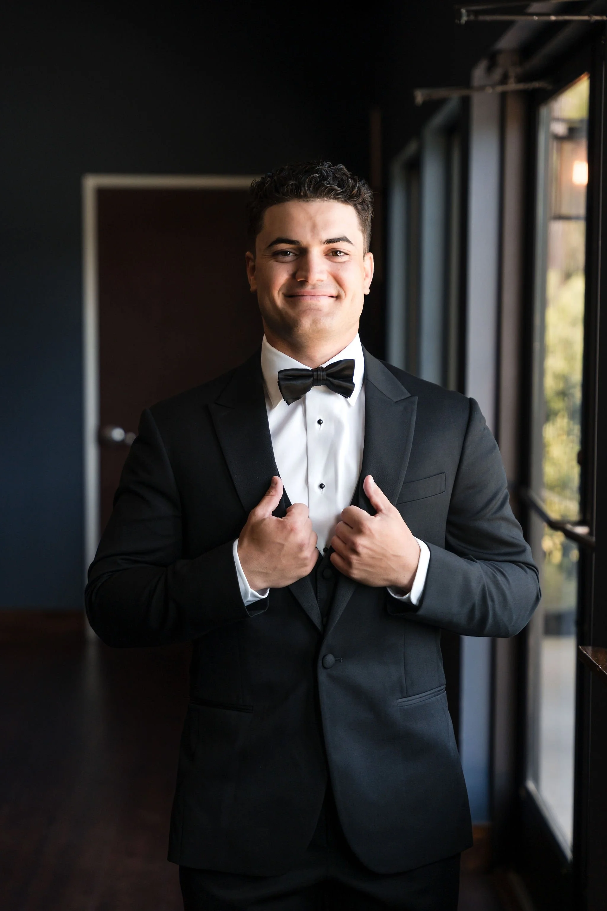 A young man dressed in a black tuxedo with a white shirt and black bow tie, standing indoors near windows, smiling and adjusting his jacket.