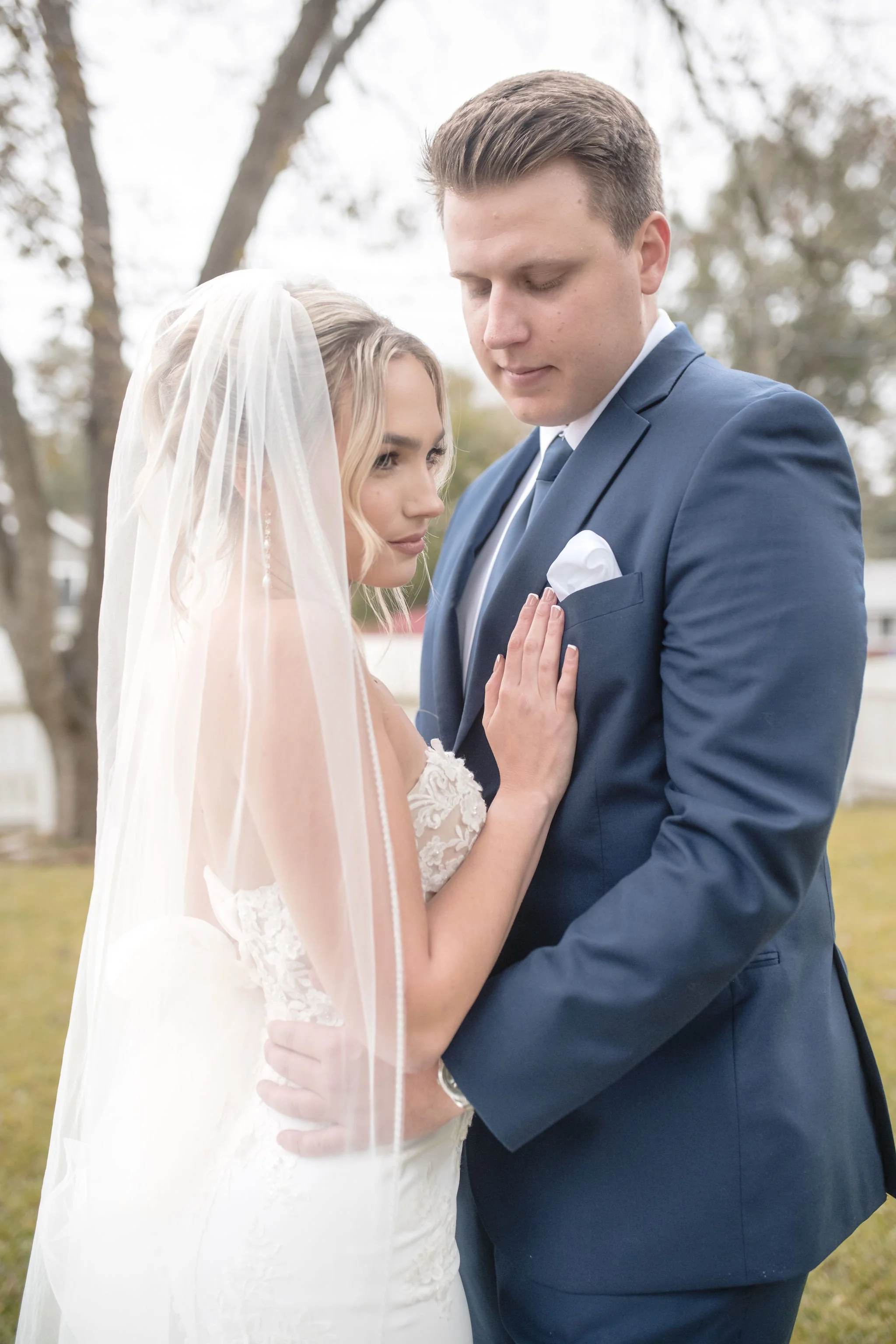 A bride and groom in wedding attire standing close outdoors, with the bride touching the groom's chest and looking at him, while the groom forms a gentle expression.