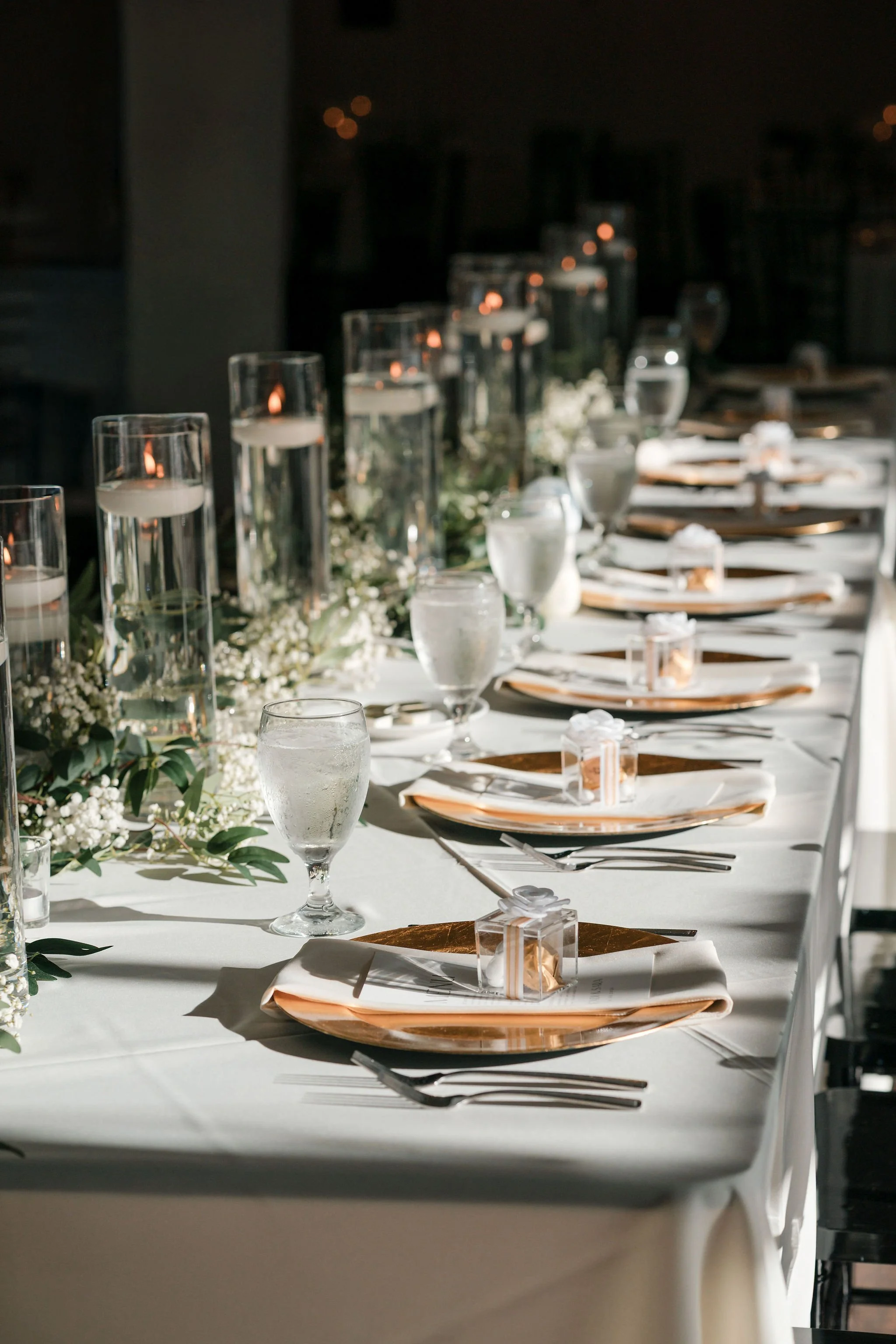 Elegant banquet table setting with white tablecloth, glasses of water, candles, floral centerpiece, and gold accents for a wedding or formal event.