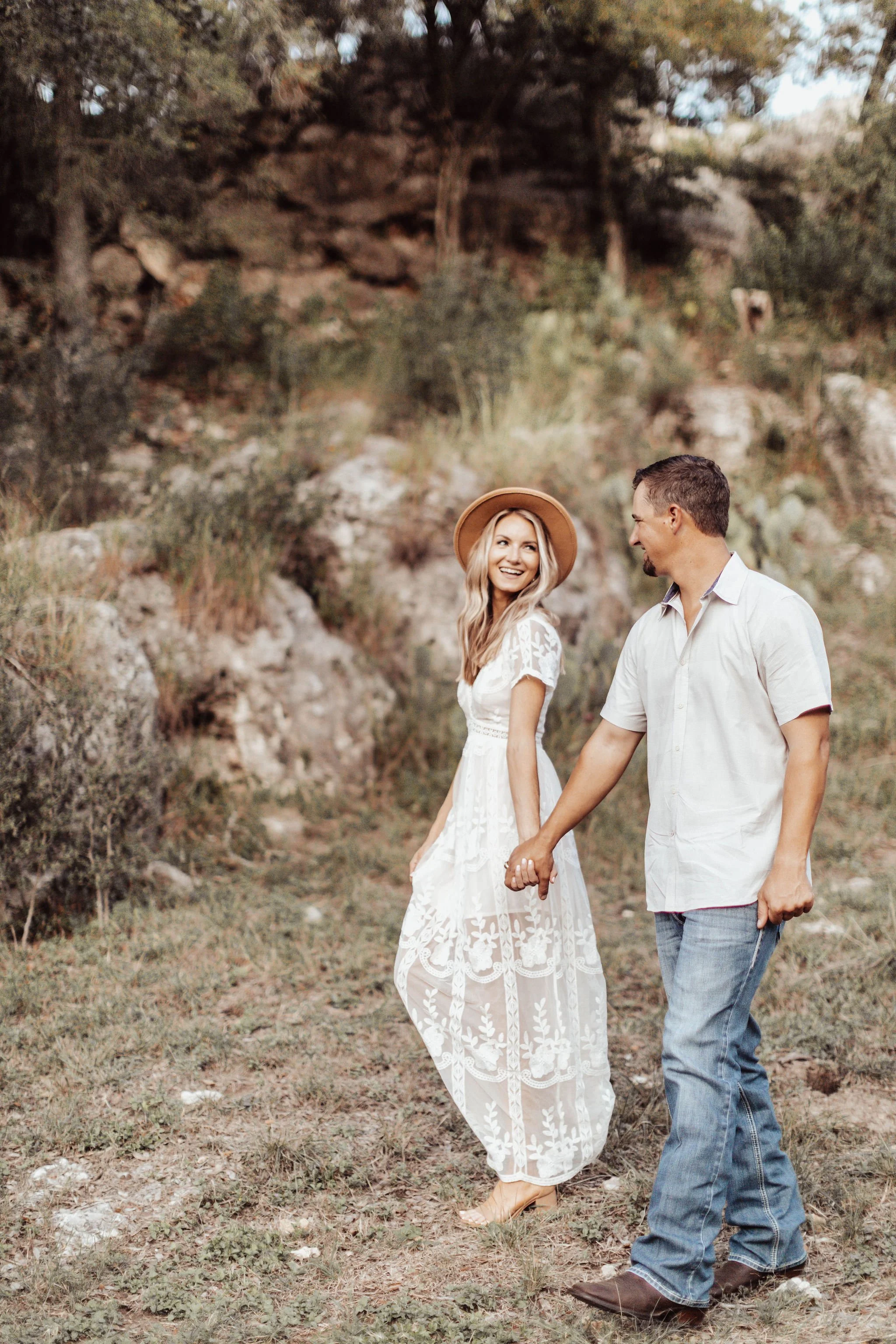 A woman wearing a white lace dress and a tan hat walking hand in hand with a man in a white shirt and jeans in a natural outdoor setting with rocks and trees.