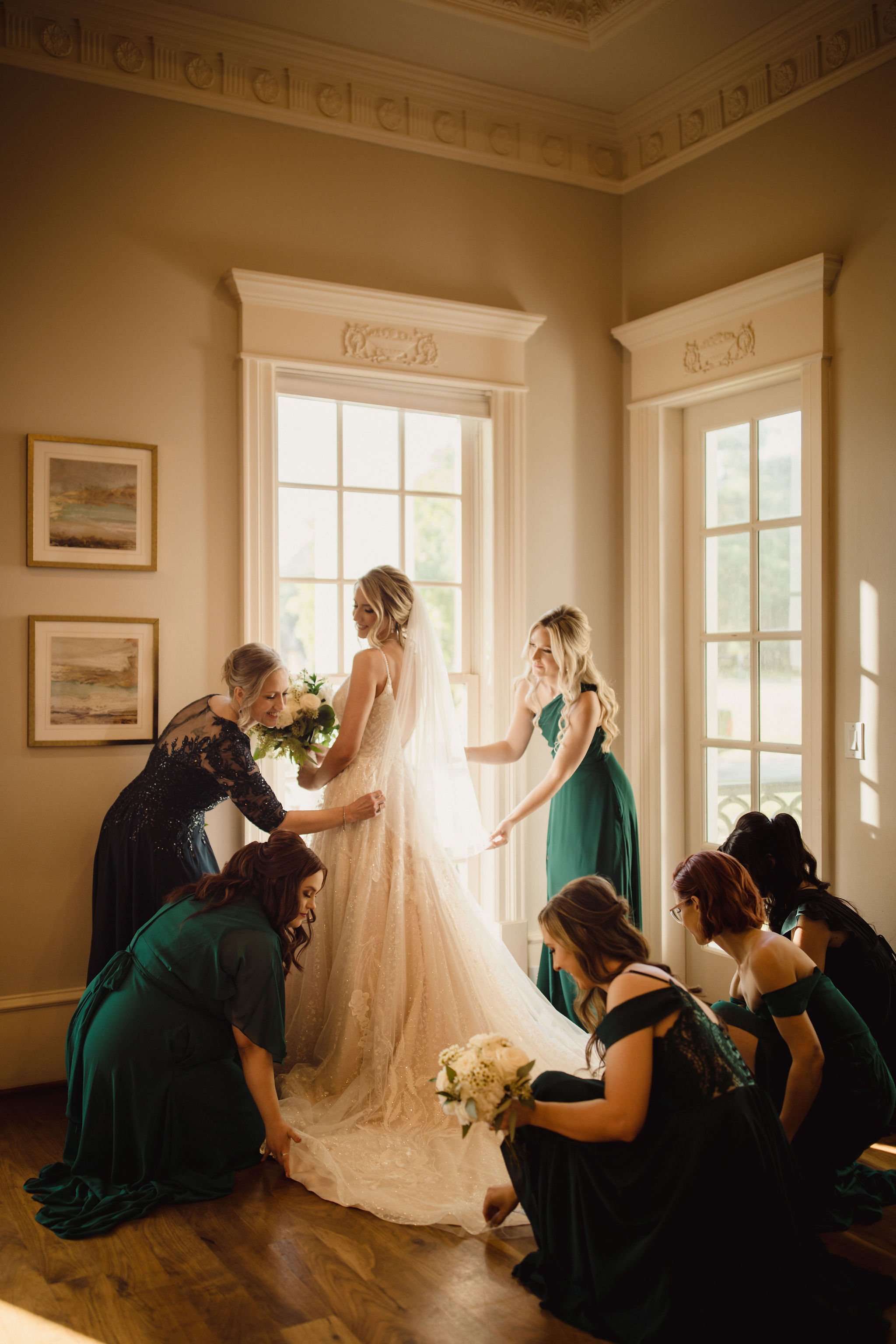 Bride in a wedding dress with seven women helping her adjust her gown in a sunlit room.