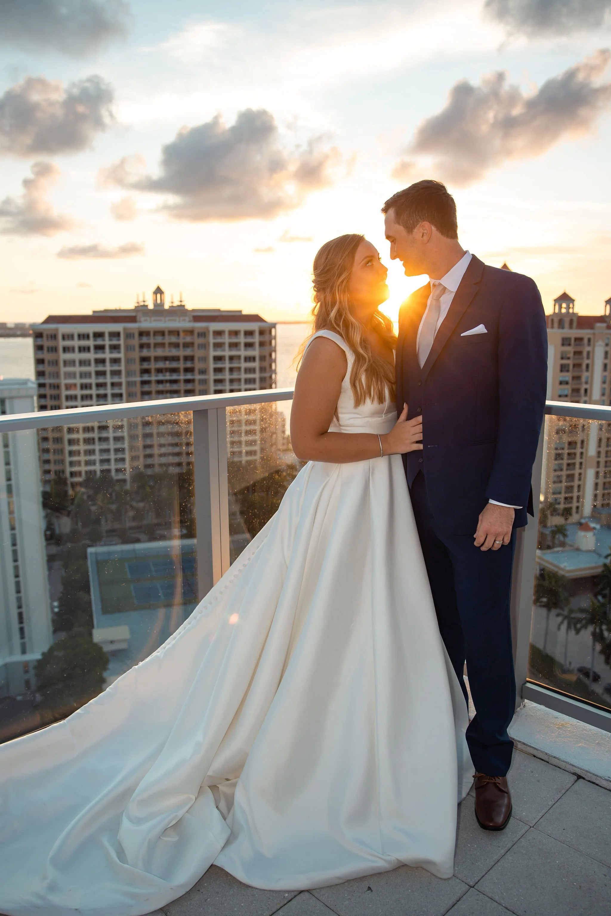 A bride and groom on a rooftop at sunset, facing each other closely, with city buildings in the background.