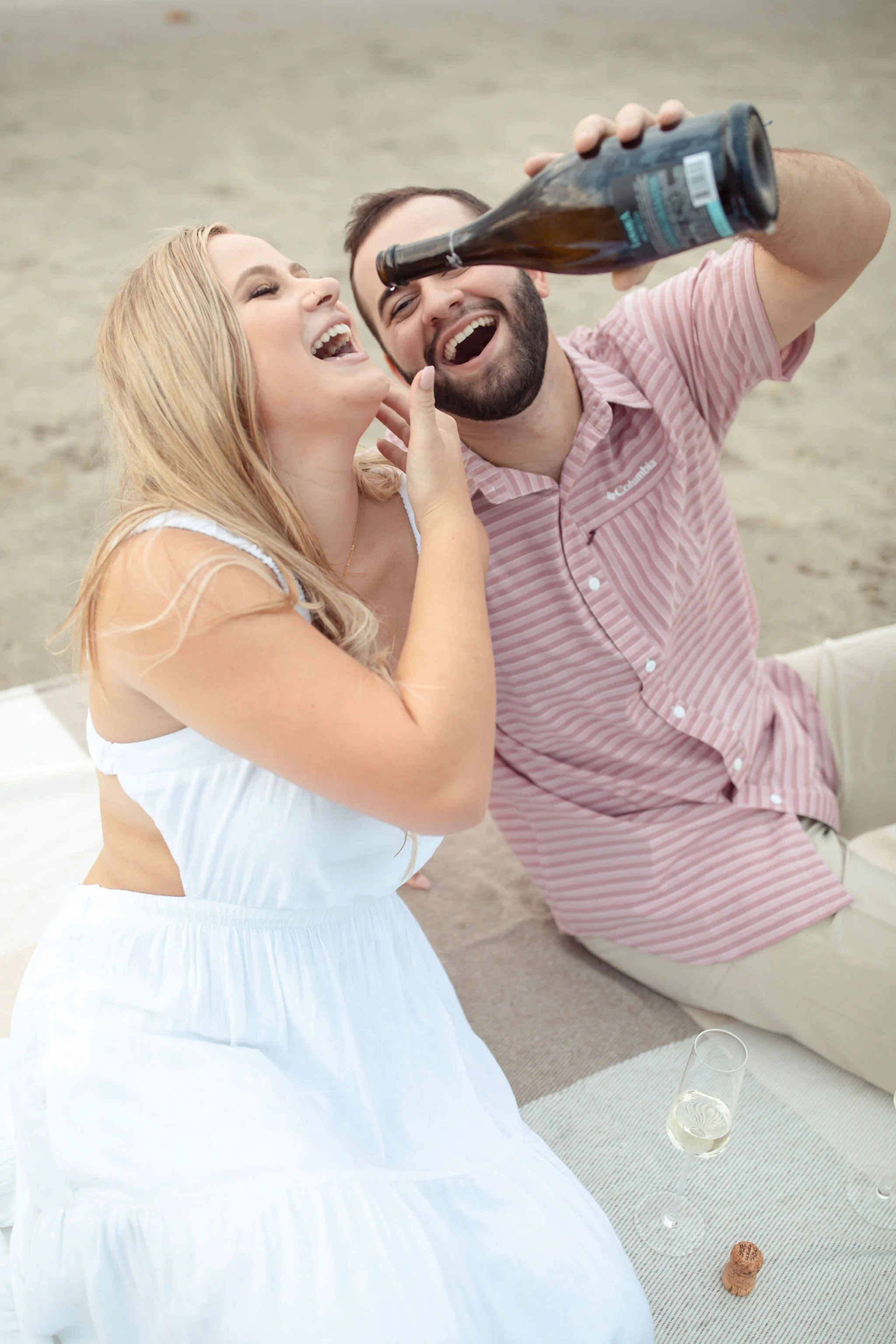 A man and woman enjoying a beach scene, with the man pouring beer into the woman's mouth, both smiling and laughing. There is a bottle of beer, a glass of champagne, and a cork on the ground.