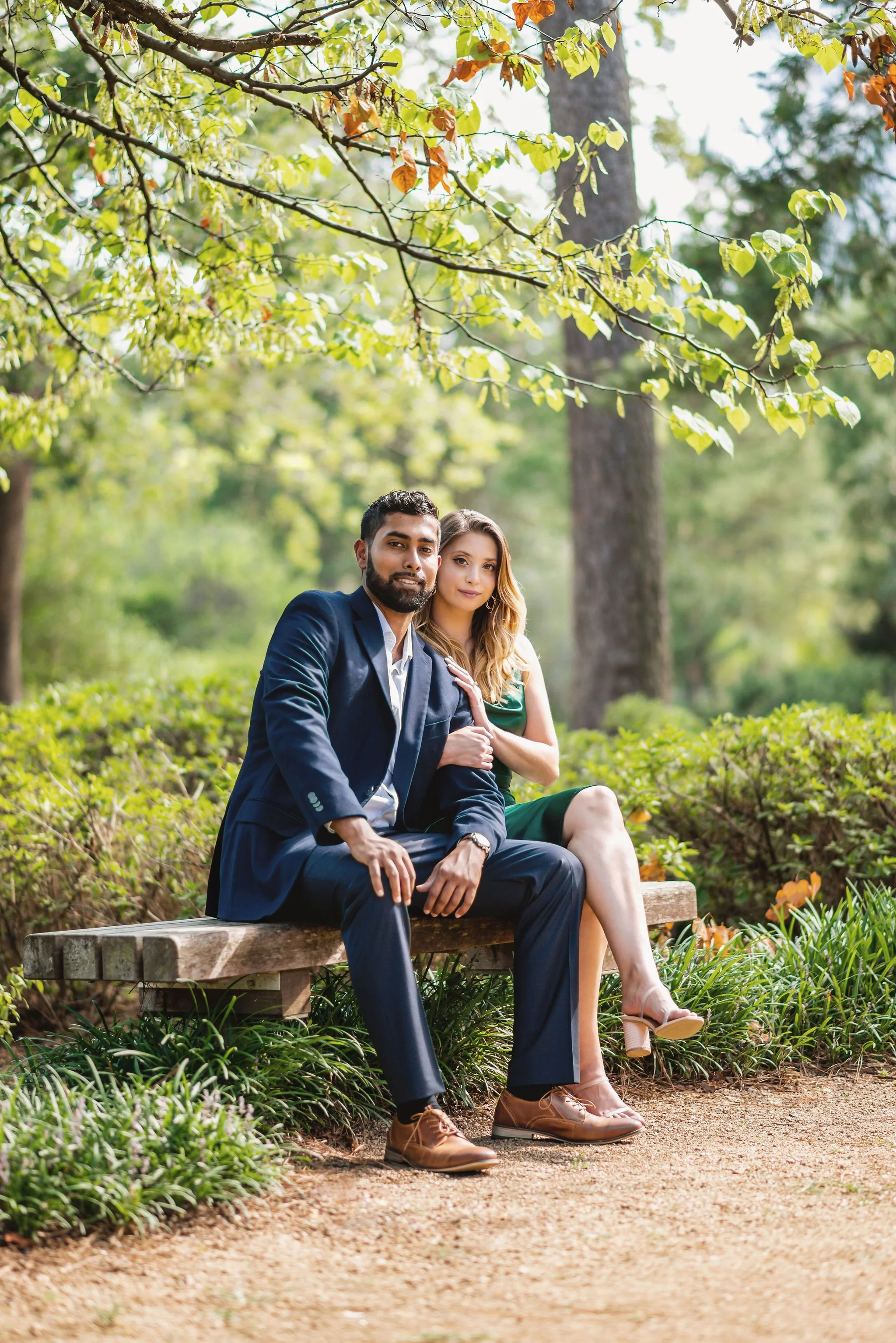 A man and woman sitting on a park bench surrounded by green trees and foliage during daytime. The man is wearing a navy blue suit, and the woman is dressed in a green dress, both looking at the camera.