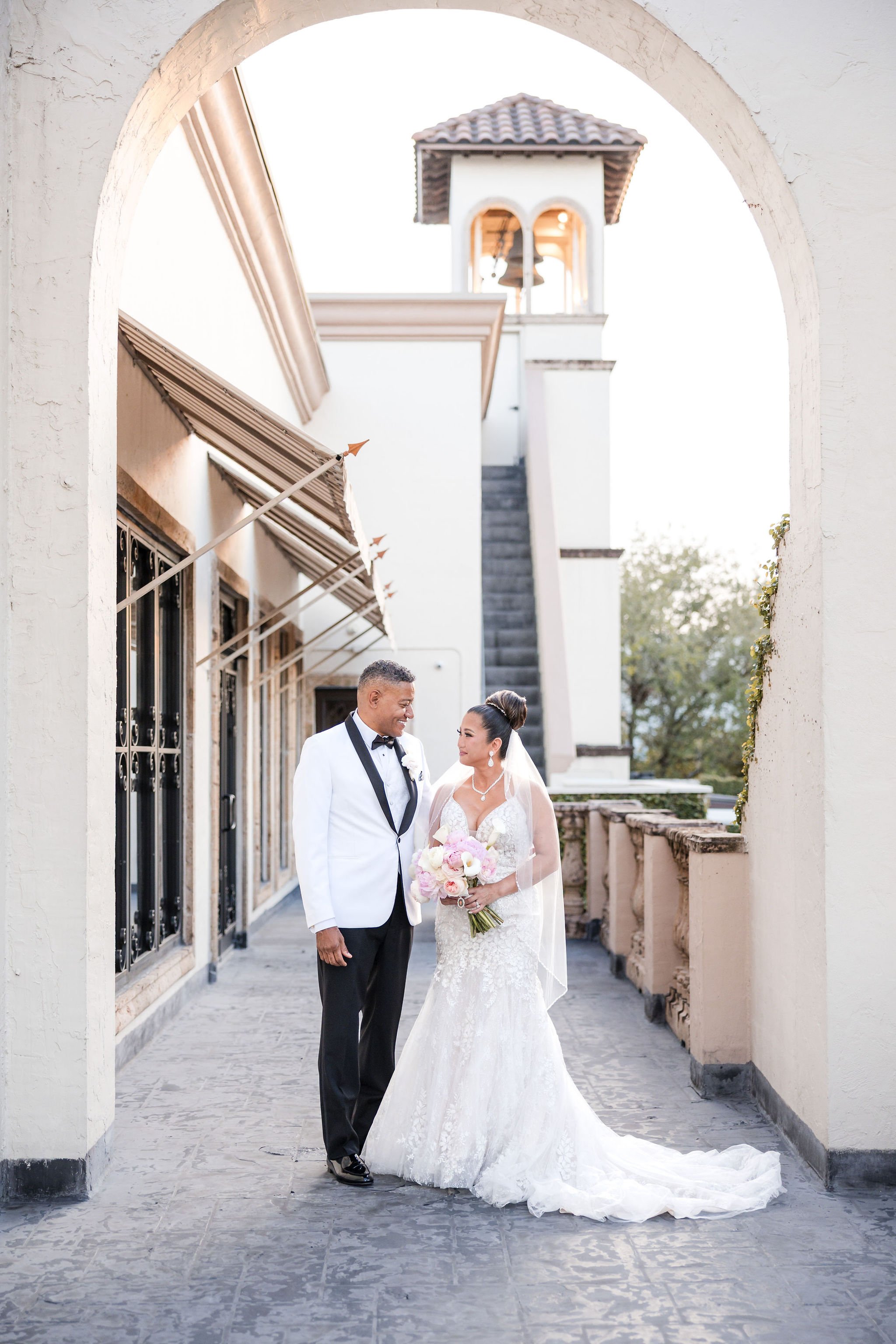 A bride and groom in wedding attire sharing a moment under an archway outside a building with a bell tower in the background.