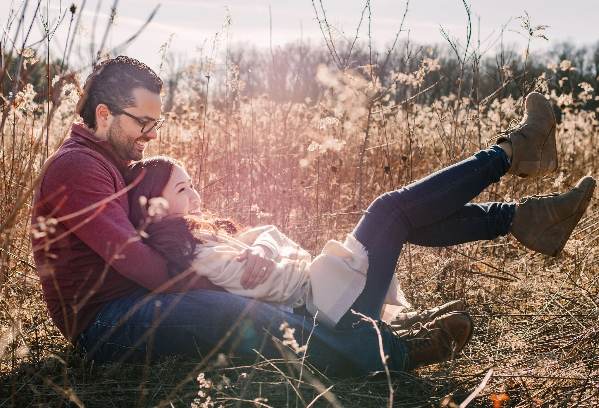 A couple sitting in a field of dry grass and wildflowers, smiling and enjoying each other's company on a sunny day.
