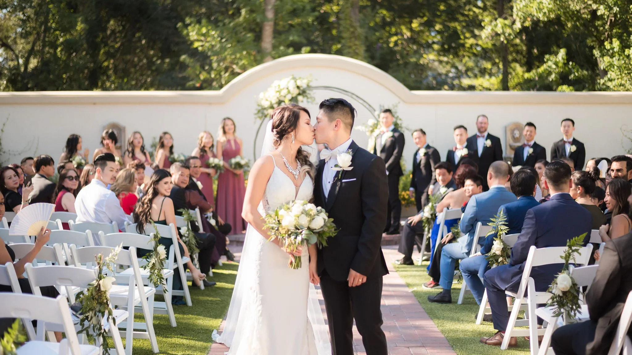 Bride and groom kissing during outdoor wedding ceremony with guests seated on white chairs, flower decorations, and a white wall backdrop.