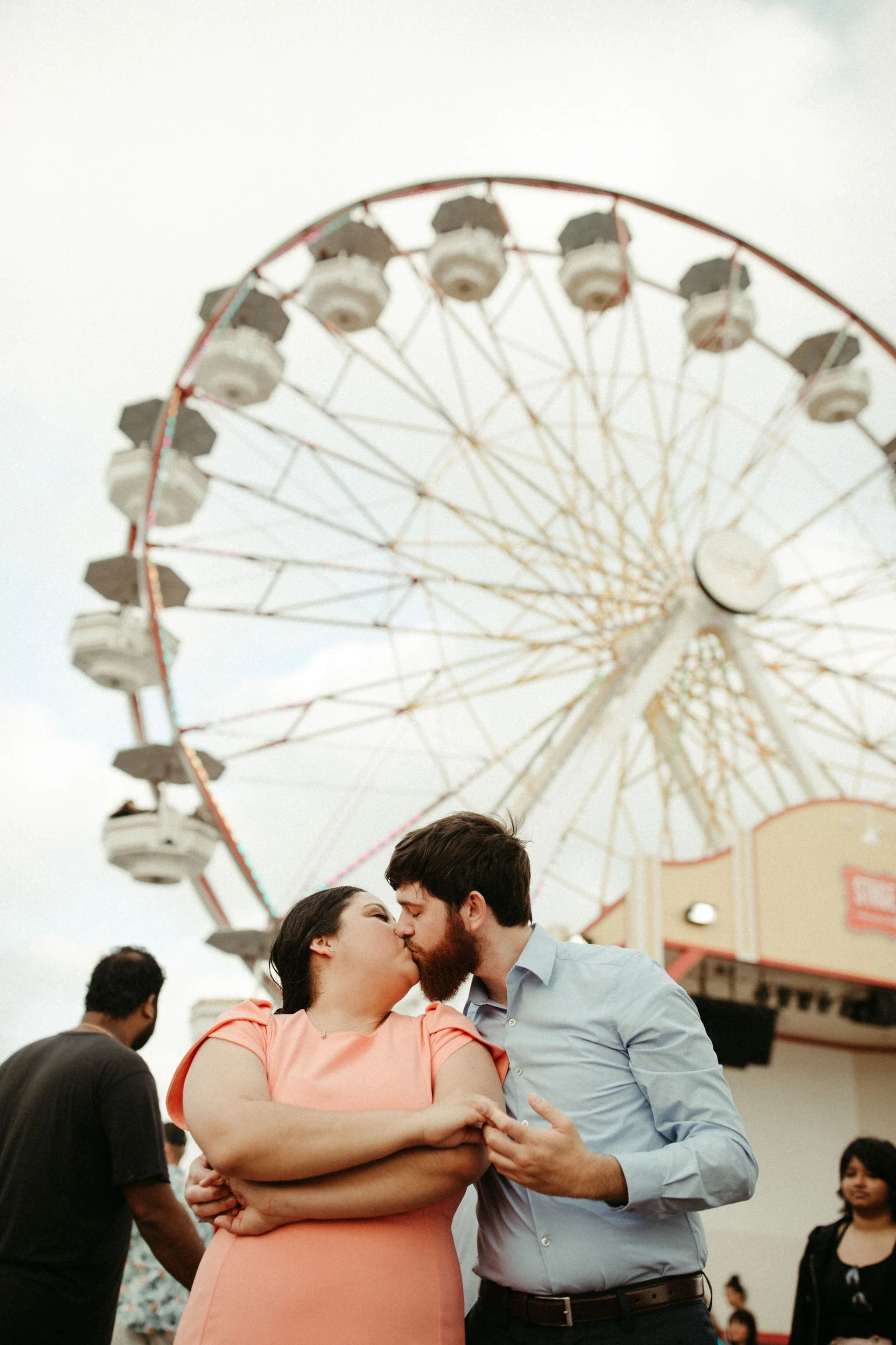 A man and woman sharing a kiss at an amusement park, with a Ferris wheel in the background and other people nearby.