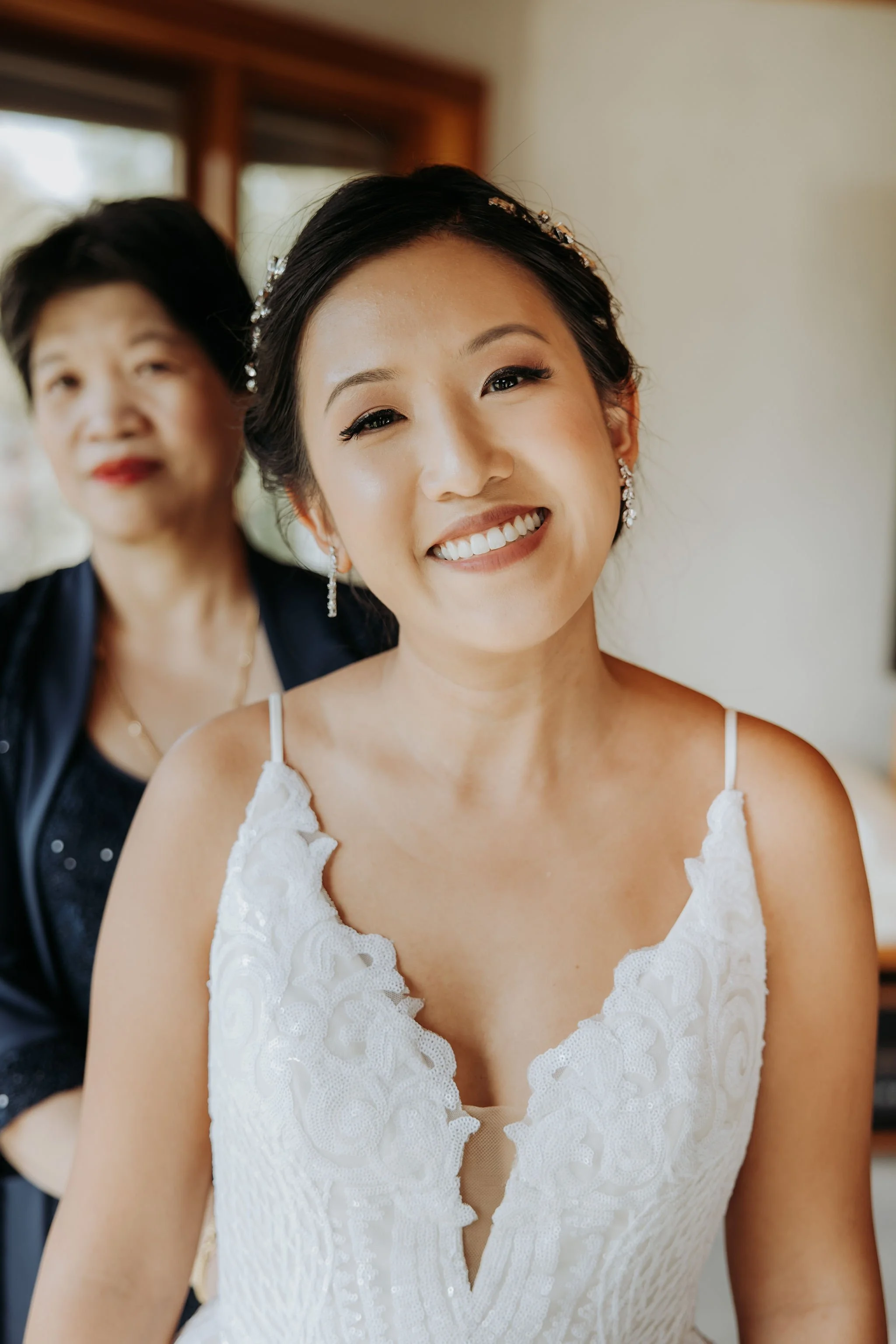 A smiling bride in a white lace wedding dress with earrings, with an older woman behind her, in a room with natural light.