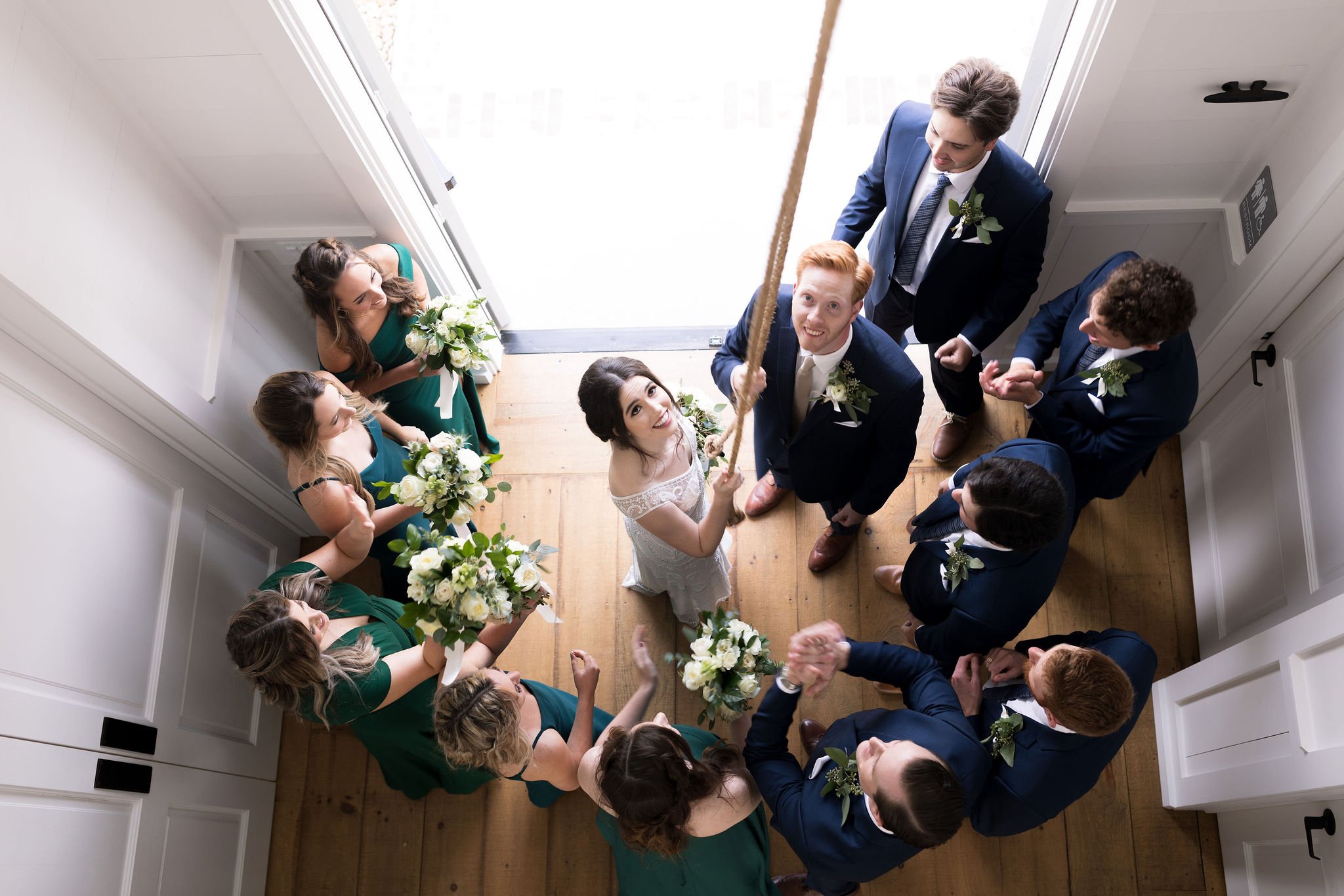 A bride and groom in a wedding party holding a rope, surrounded by bridesmaids and groomsmen in an overhead shot.