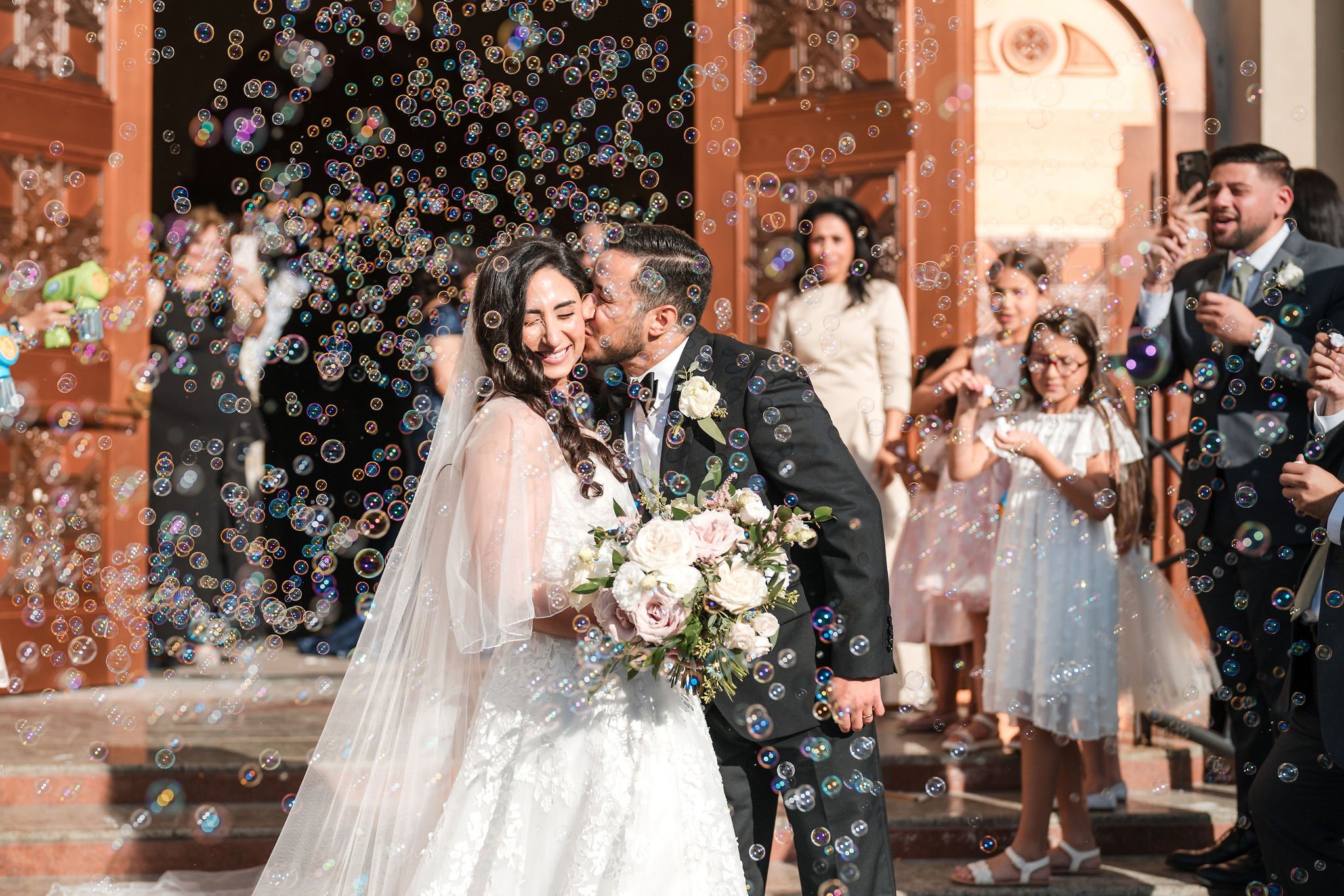 A newlywed couple shares a kiss on their wedding day surrounded by balloons and friends outside a ceremonial building.