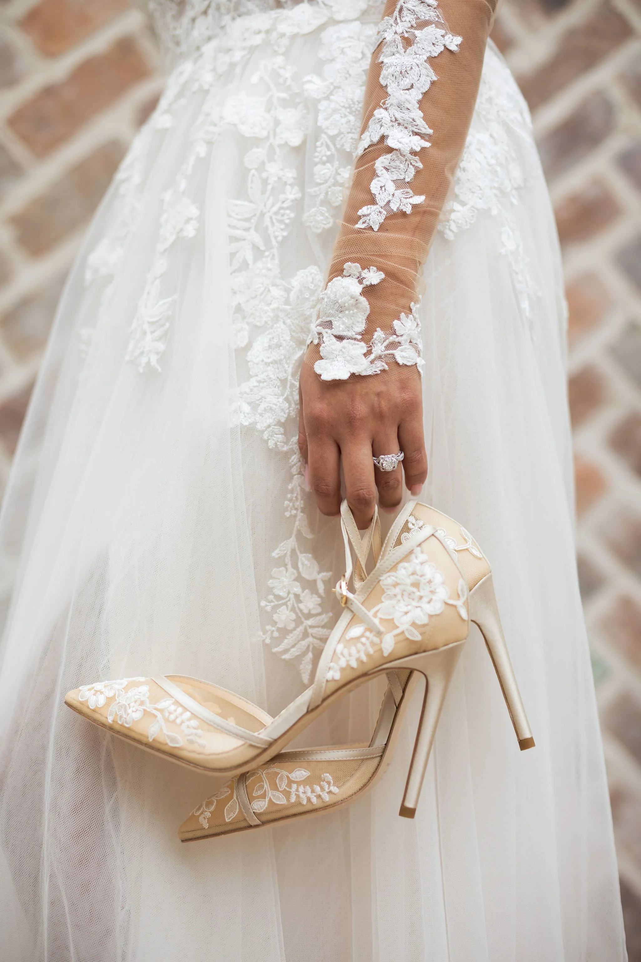 Close-up of a bride in a lace wedding dress holding a beige high heel shoe with intricate floral embroidery.