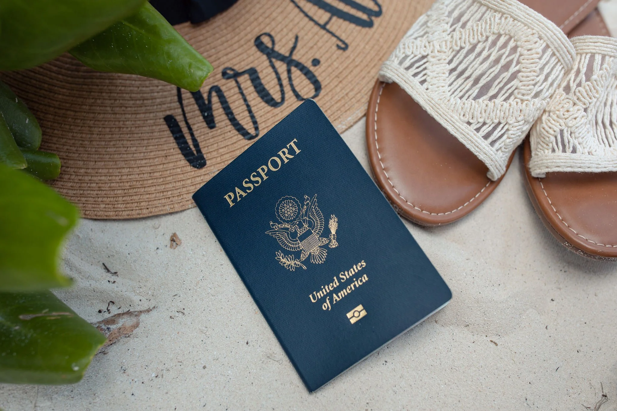 United States passport on sandy surface next to beige woven sandals, a straw hat, and green plants.