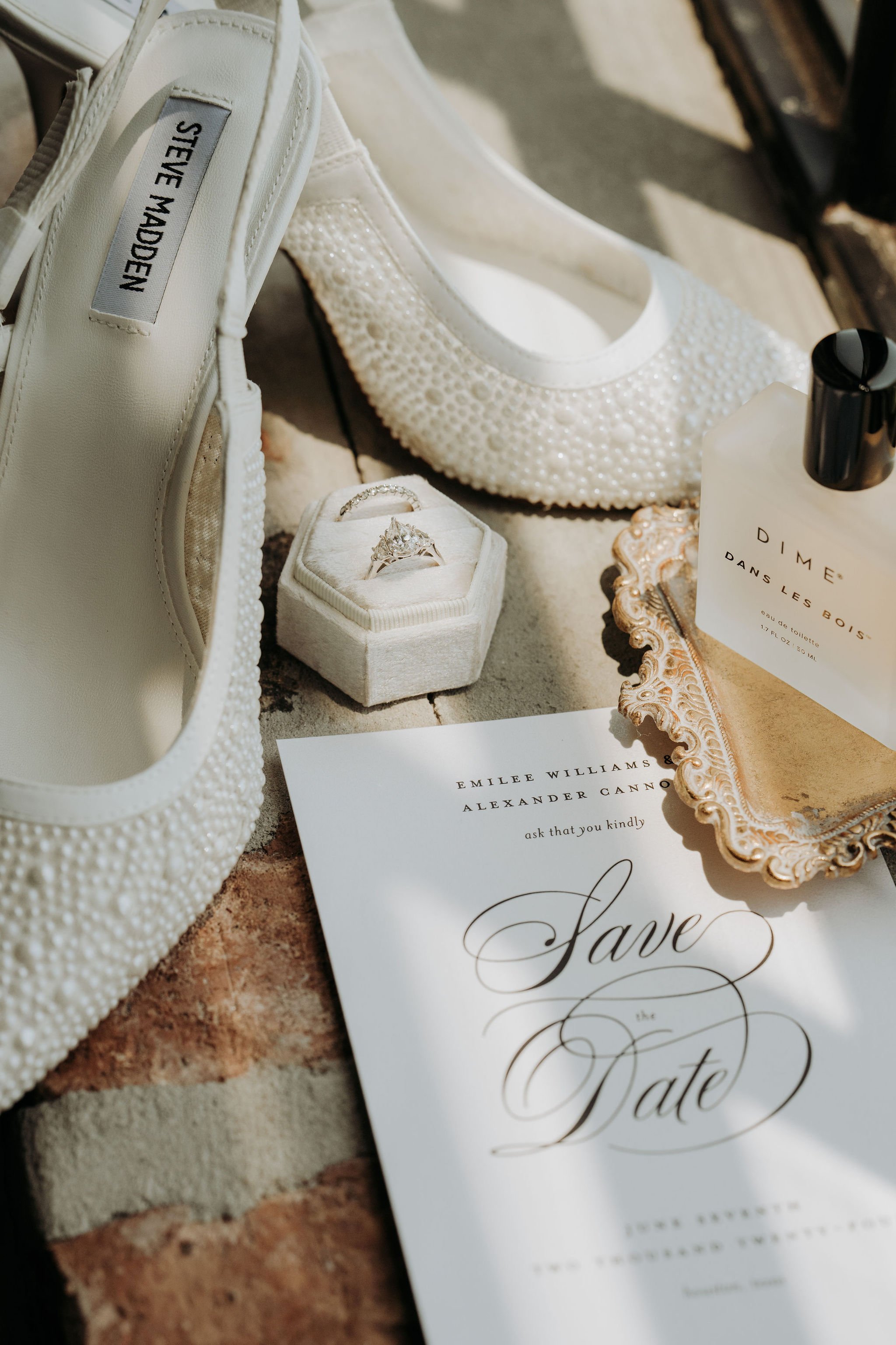 Wedding items including a white Steve Madden shoe with pearl embellishments, a ring in a white box, a drinking bottle labeled 'DIME DANS LES BOIS,' a gold decorative tray, and a save the date card for Emily Williams and Alexander Cannon.