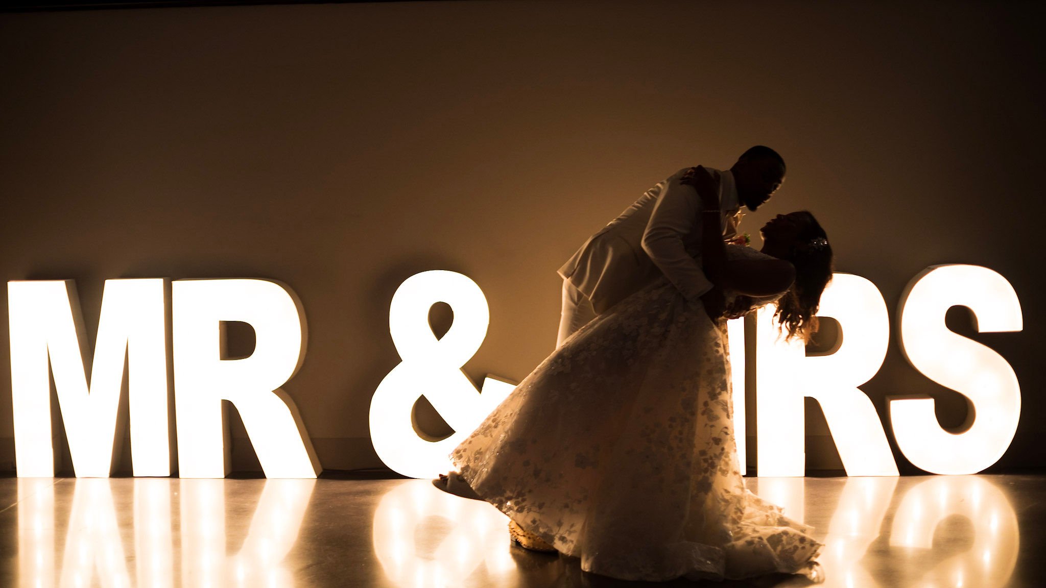 A couple, dressed in wedding attire, sharing a kiss during their wedding reception, silhouetted against large illuminated letters spelling 'MR & MRS'.