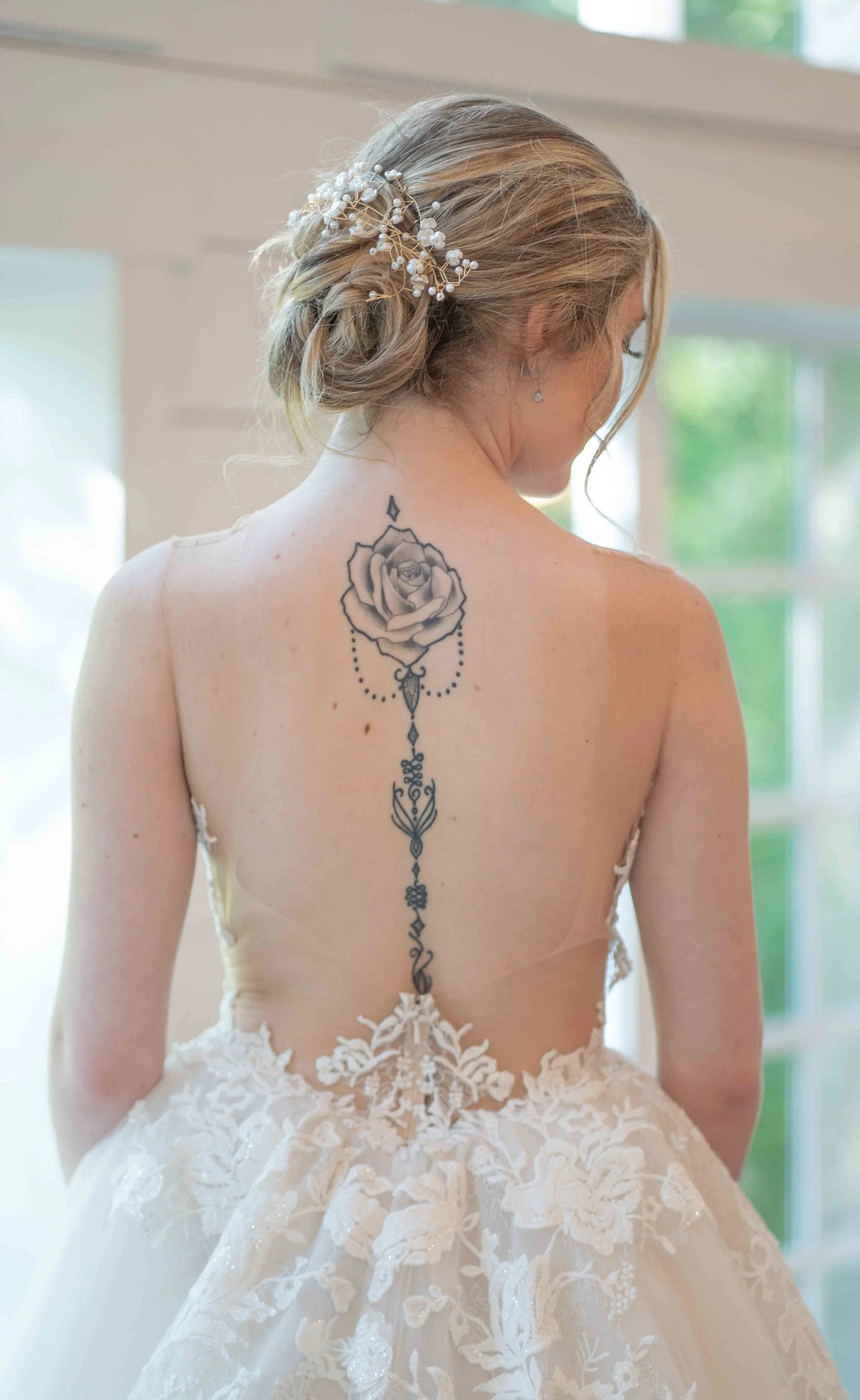 A woman with blonde hair styled in loose updo with a pearl and floral hairpiece, showing her back with a large tattoo of a rose and decorative designs running down her spine, wearing a wedding dress with lace and floral embroidery.