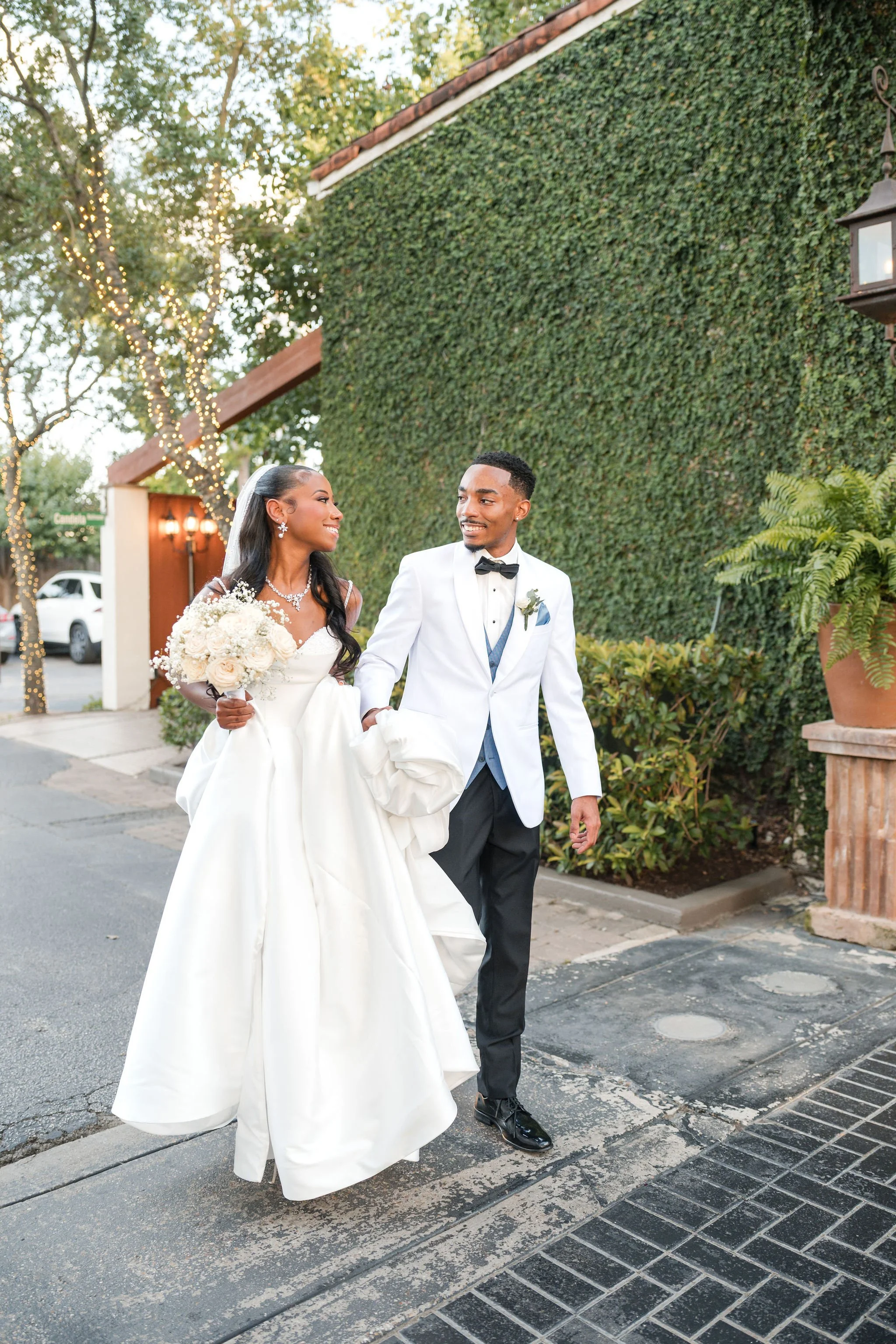 A newlywed couple, a woman in a white wedding gown holding a bouquet and a man in a white tuxedo jacket with black pants, walking outside on a sidewalk during daytime, with a backdrop of green leafy wall and trees decorated with string lights.