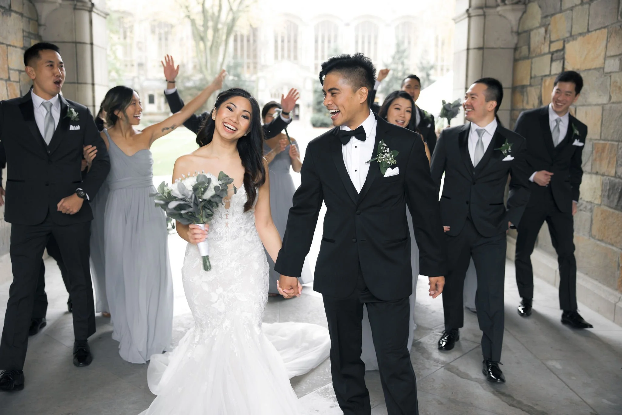 Happy bride and groom walking hand in hand, surrounded by their wedding party, celebrating outdoors.