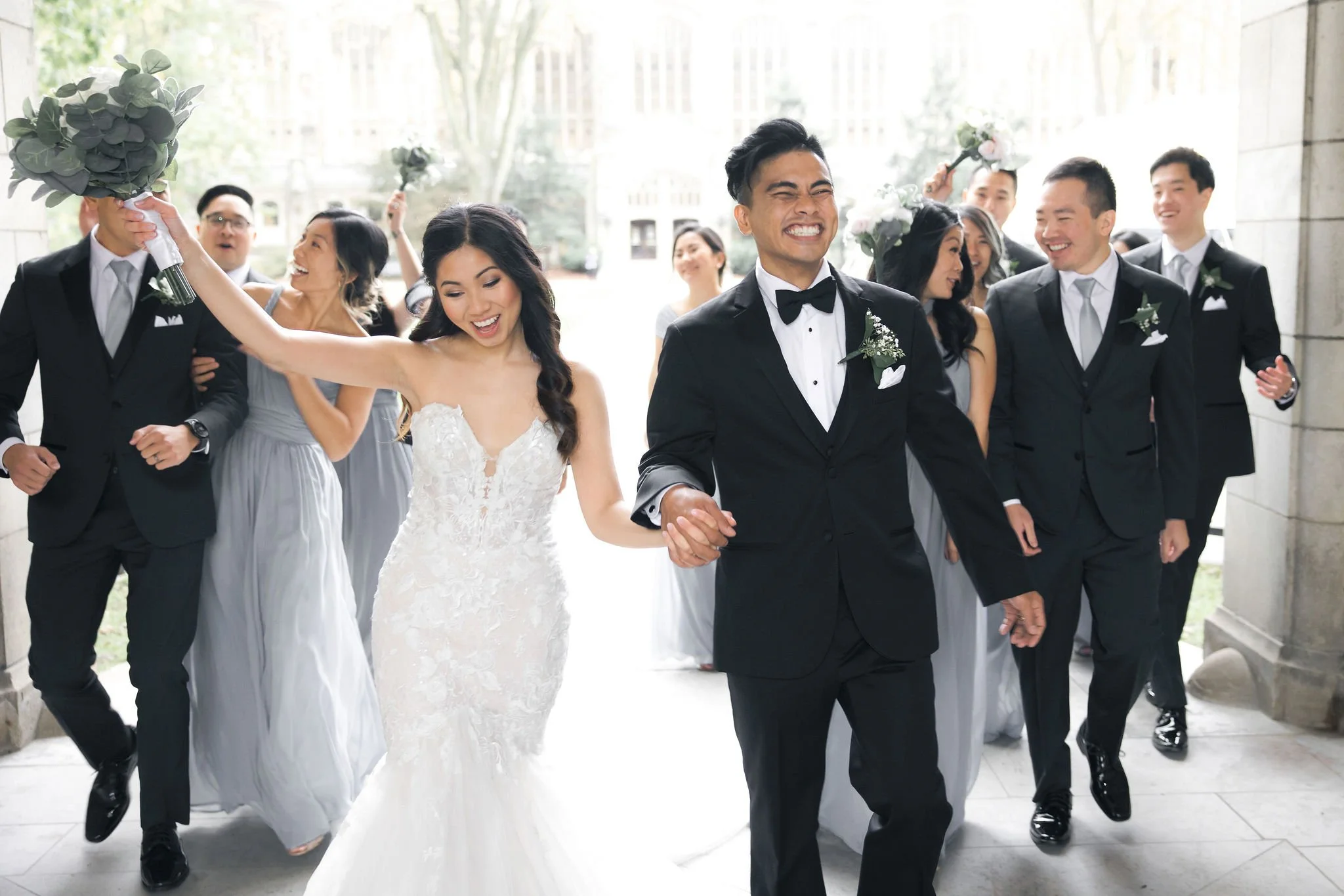 A wedding celebration with the bride and groom leading friends outside, the bride in a white wedding dress holding a bouquet, the groom in a black tuxedo, everyone smiling, some carrying flowers, sunny outdoor setting.