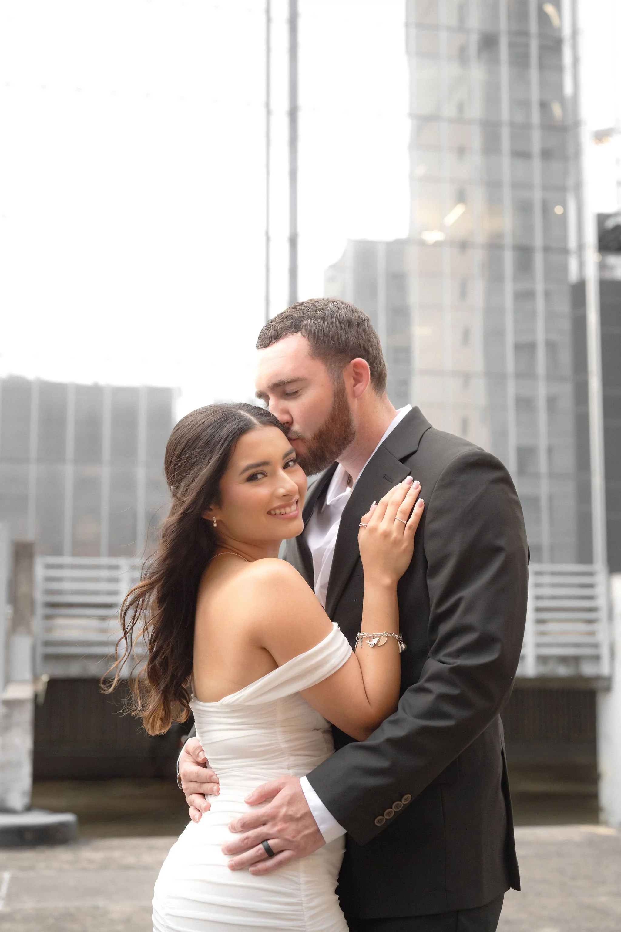 A newlywed couple embracing in an urban setting, with the groom kissing the bride on her forehead and the bride smiling.
