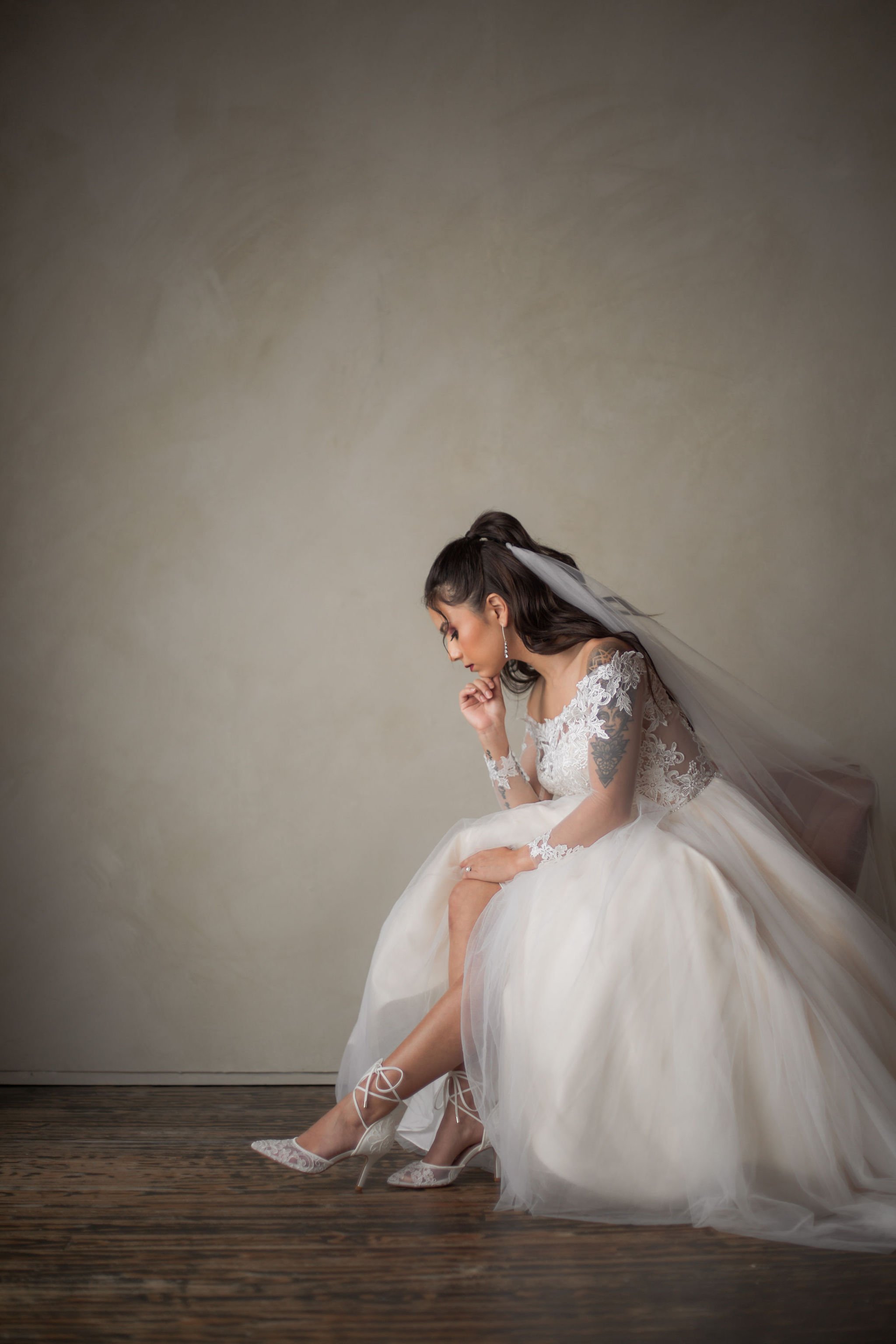 A bride in a white wedding dress sitting on a wooden bench, adjusting her high-heeled shoes, with a contemplative expression.