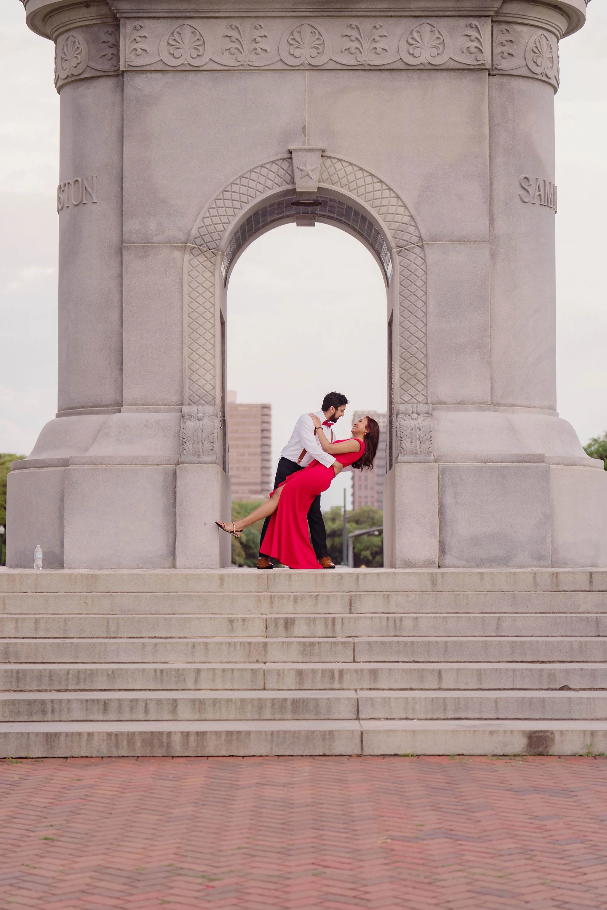 A couple dancing beneath a large stone arch monument, with a man in a white shirt and dark pants holding a woman in a red dress. The monument has decorative carvings and a star above the arch, with steps leading up to it and a cityscape in the backgr