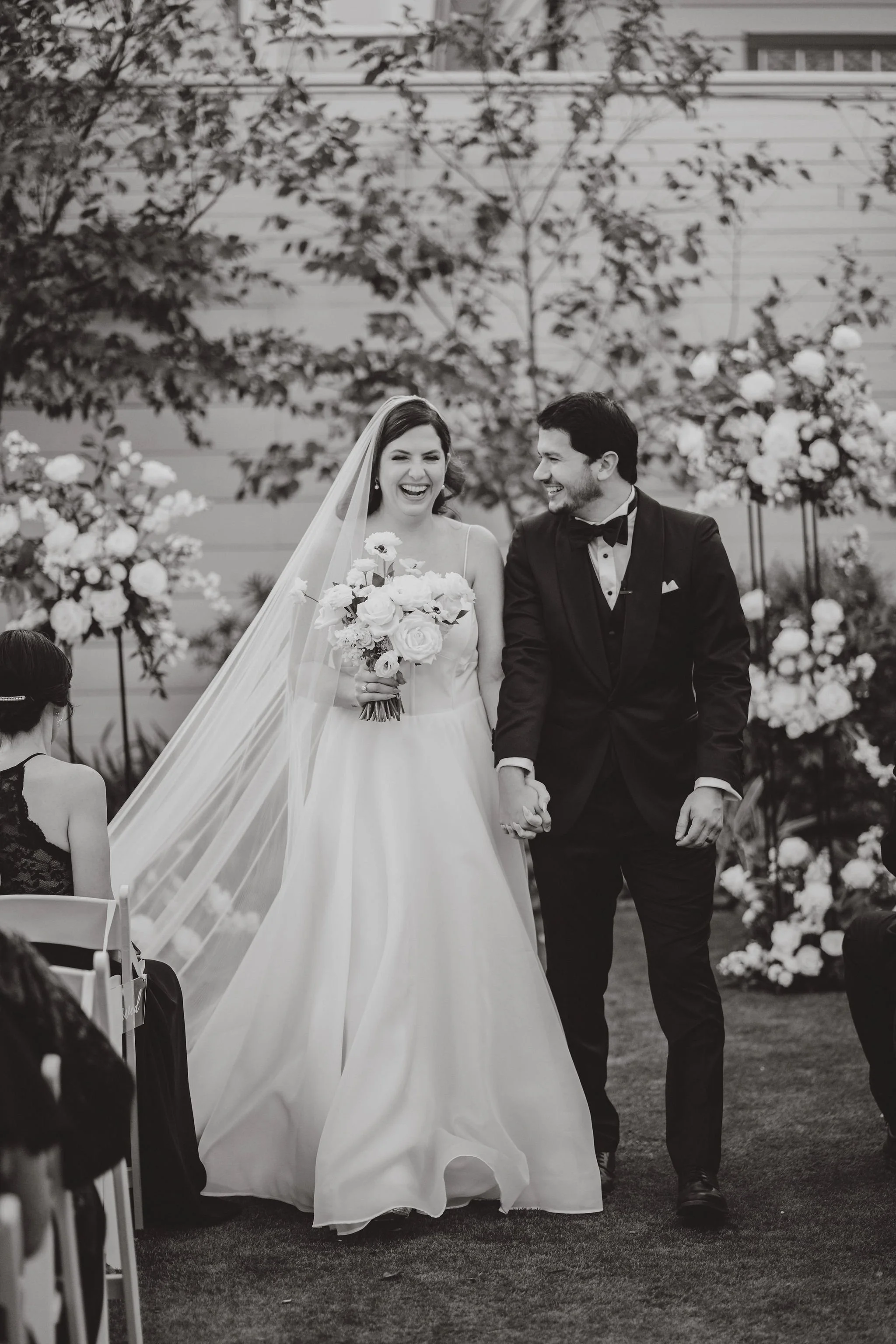 A newly married couple, the bride wearing a wedding dress and veil holding a bouquet, and the groom in a tuxedo, walking hand in hand and smiling at each other during their wedding ceremony outdoors with floral decorations.