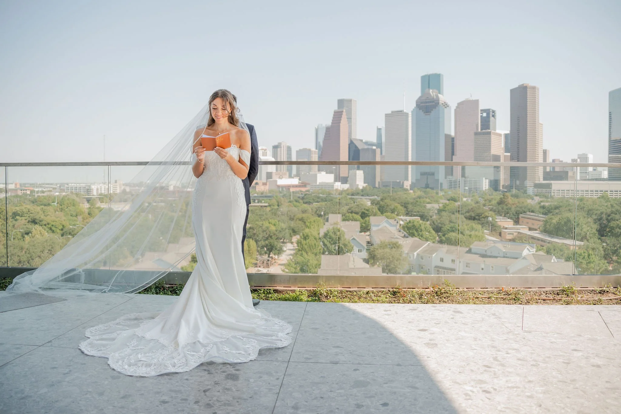 Bride in a white wedding dress with long train and veil, holding a small book, standing on a rooftop terrace with a city skyline in the background.