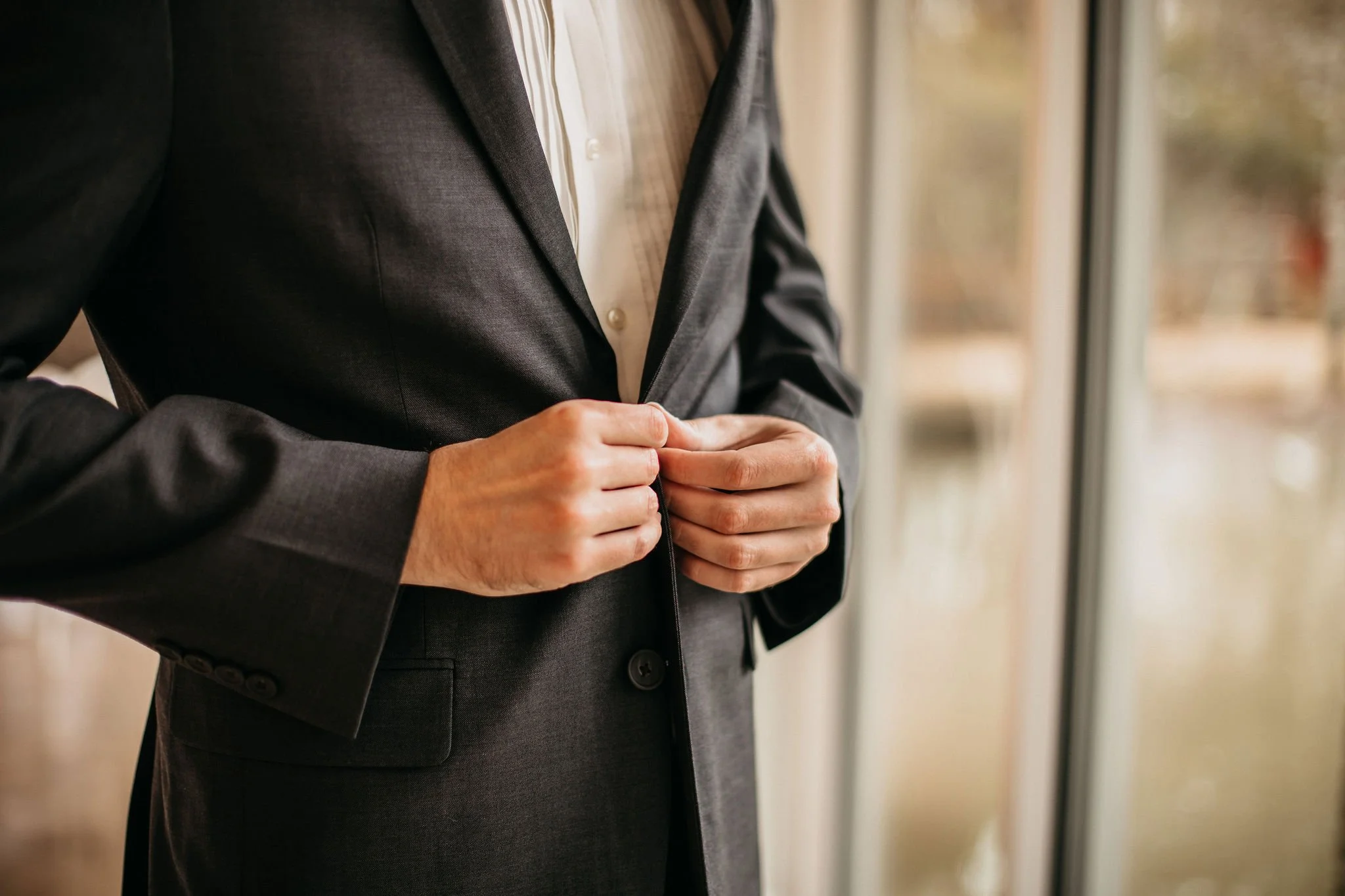 Close-up of a person in a dark suit buttoning the jacket, standing by a window with a blurred outdoor background.
