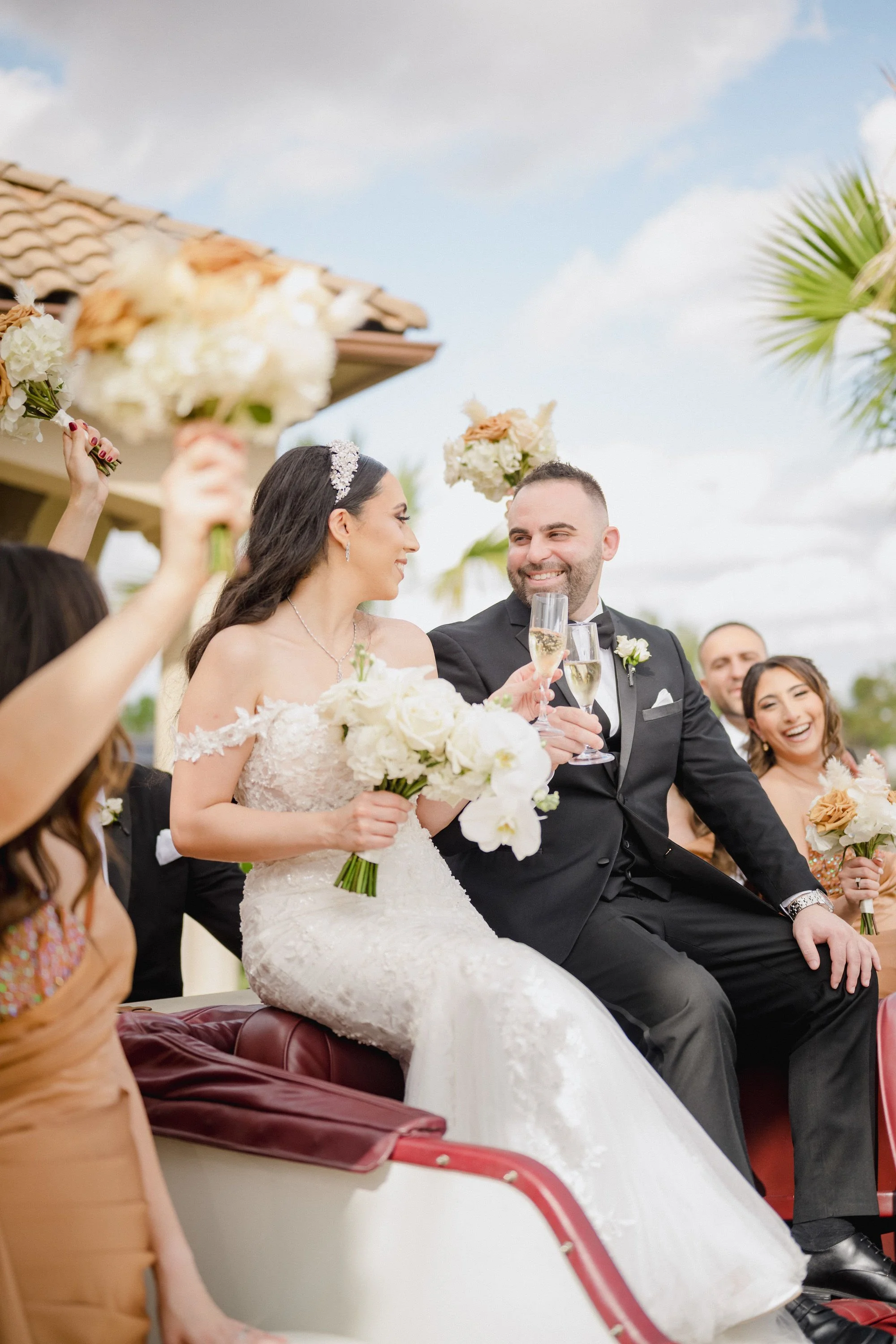 Bride and groom celebrating with friends during a parade, seated on a convertible car, holding champagne glasses, with the bride holding a bouquet of white flowers, everyone smiling and raising their glasses.