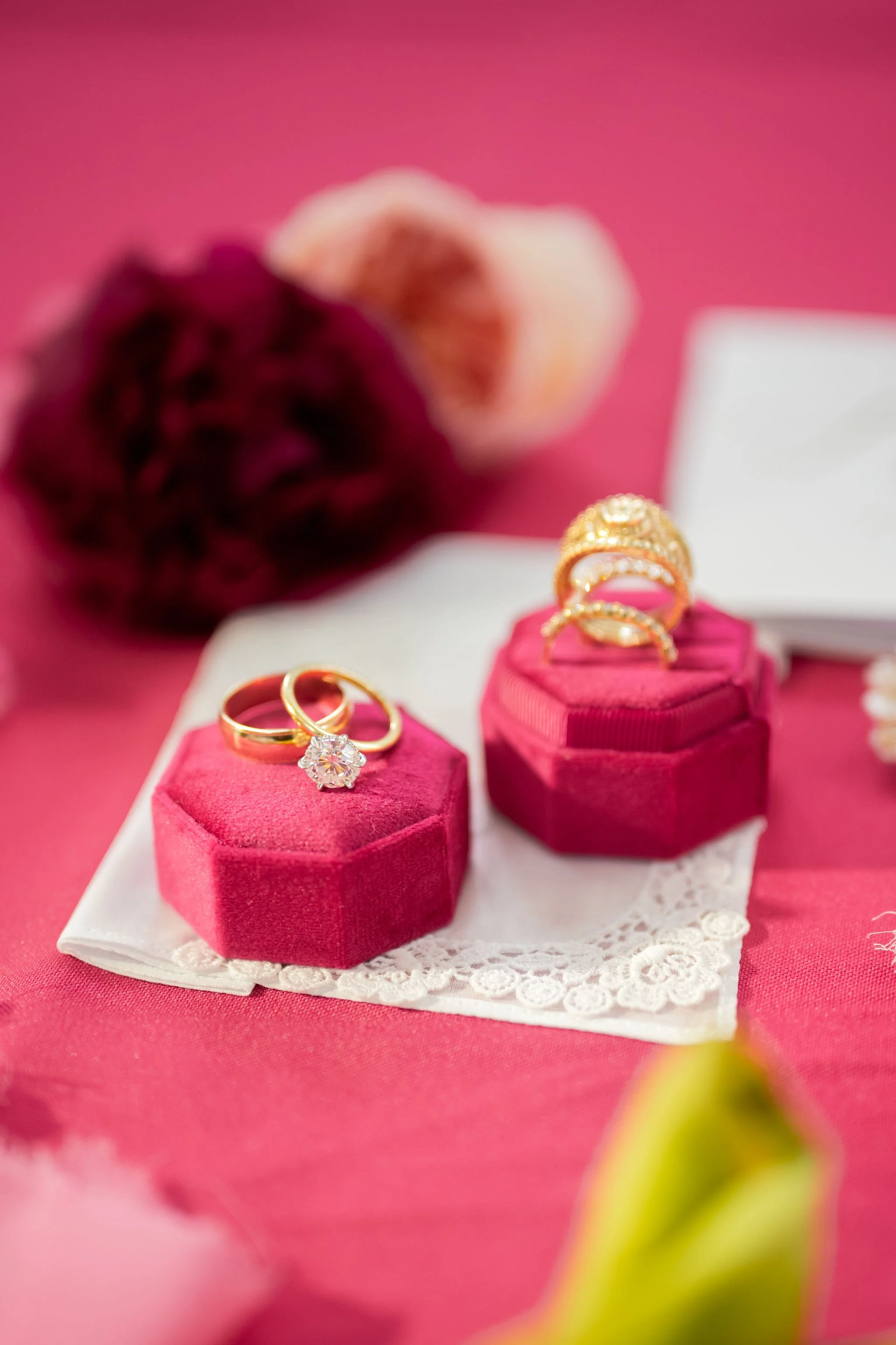 Gold rings with gemstones displayed on pink velvet ring boxes on a lace doily, with pink and purple flowers in the blurred background.