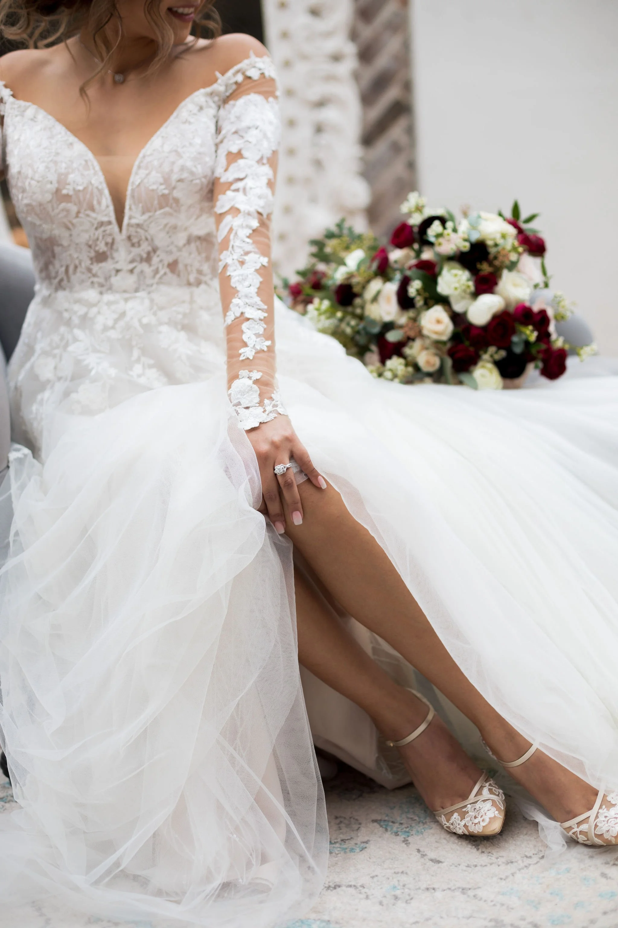 Bride in a lace wedding dress adjusting her shoe, with a bouquet of flowers nearby.