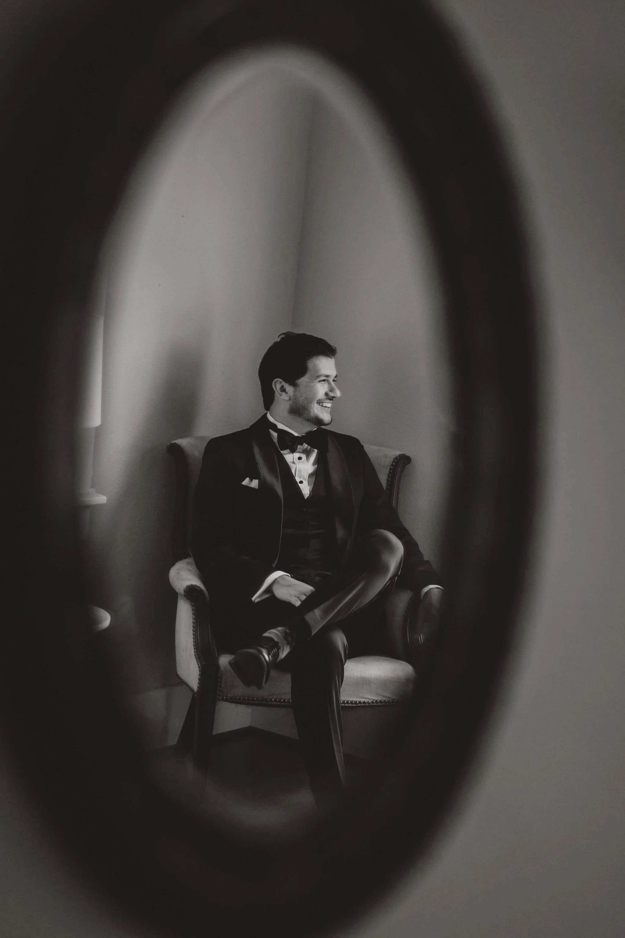 A black-and-white photo of a man in a tuxedo sitting on an armchair, framed through an oval-shaped mirror. The man is smiling and looking to his left.