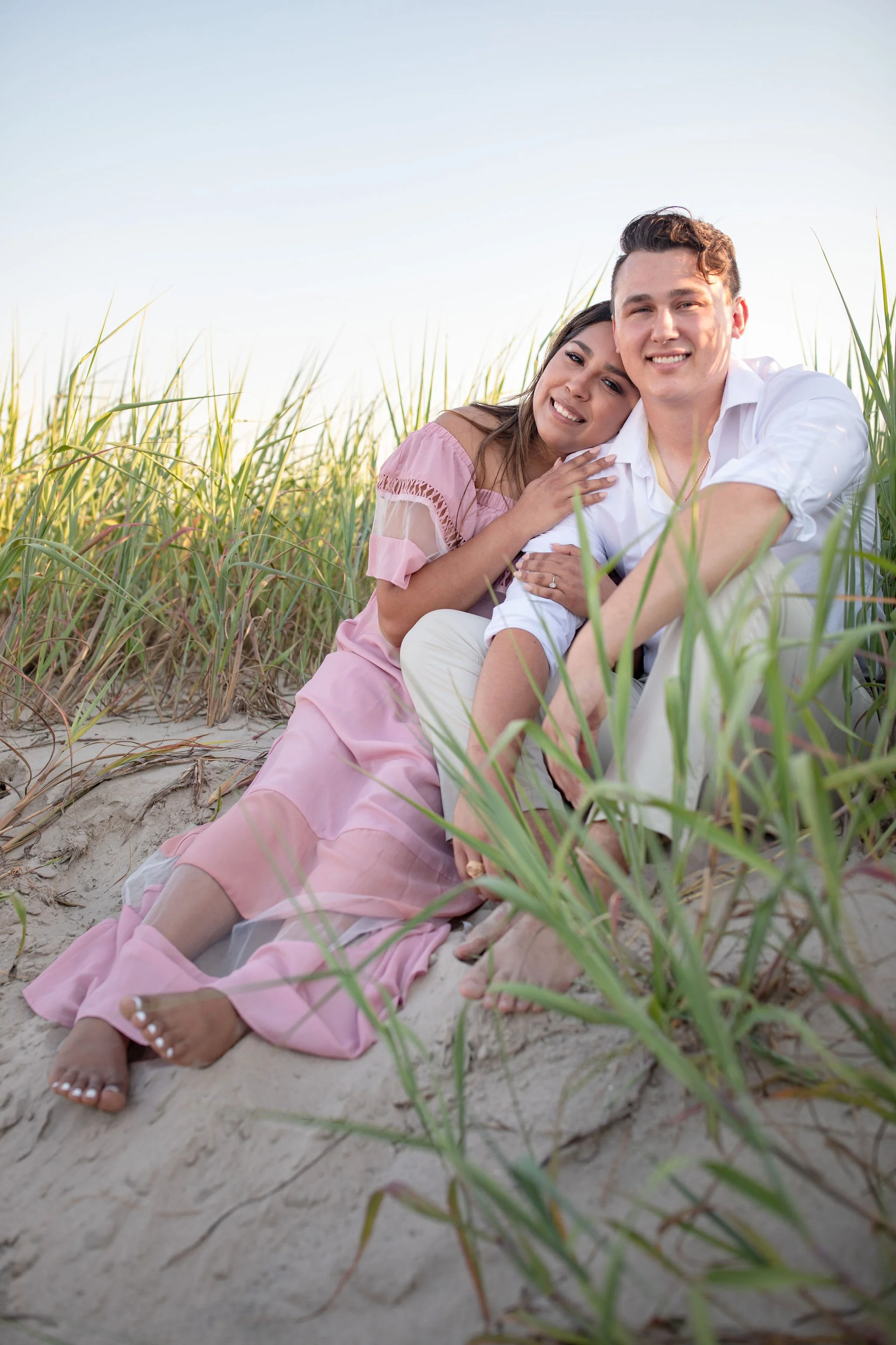 A smiling couple sitting on sandy ground among green grass, embracing each other on a bright day.
