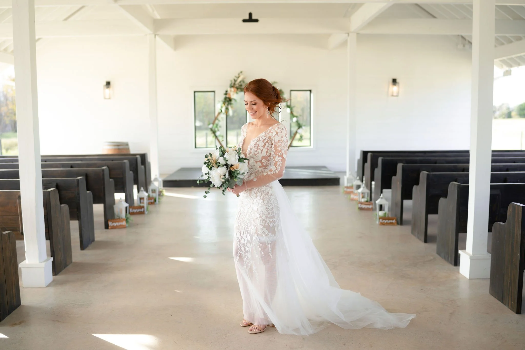 A bride in a lace wedding dress holding a bouquet of white roses, standing inside a decorated wedding venue with black benches and a floral arch in the background.