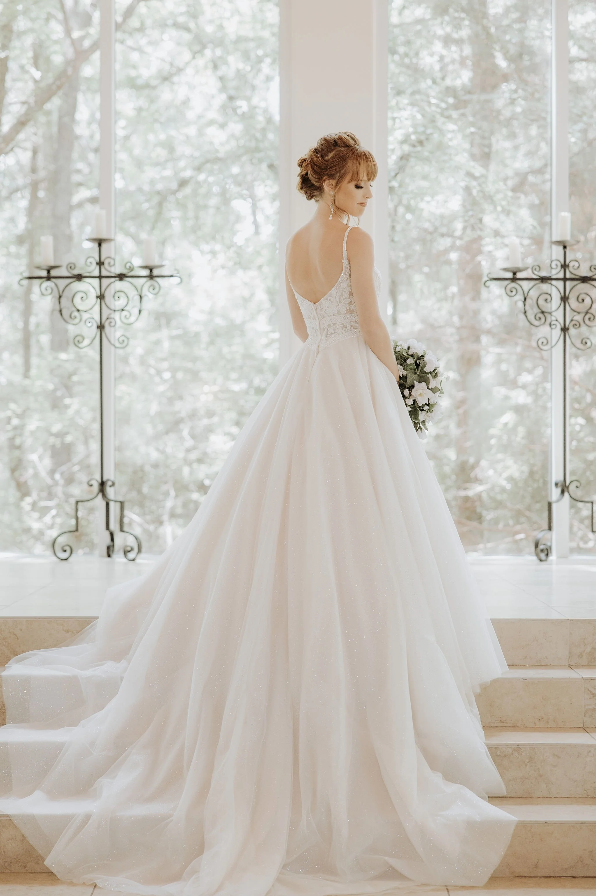 A bride in a wedding dress standing on stairs indoors with large windows and trees outside, holding a bouquet of flowers.