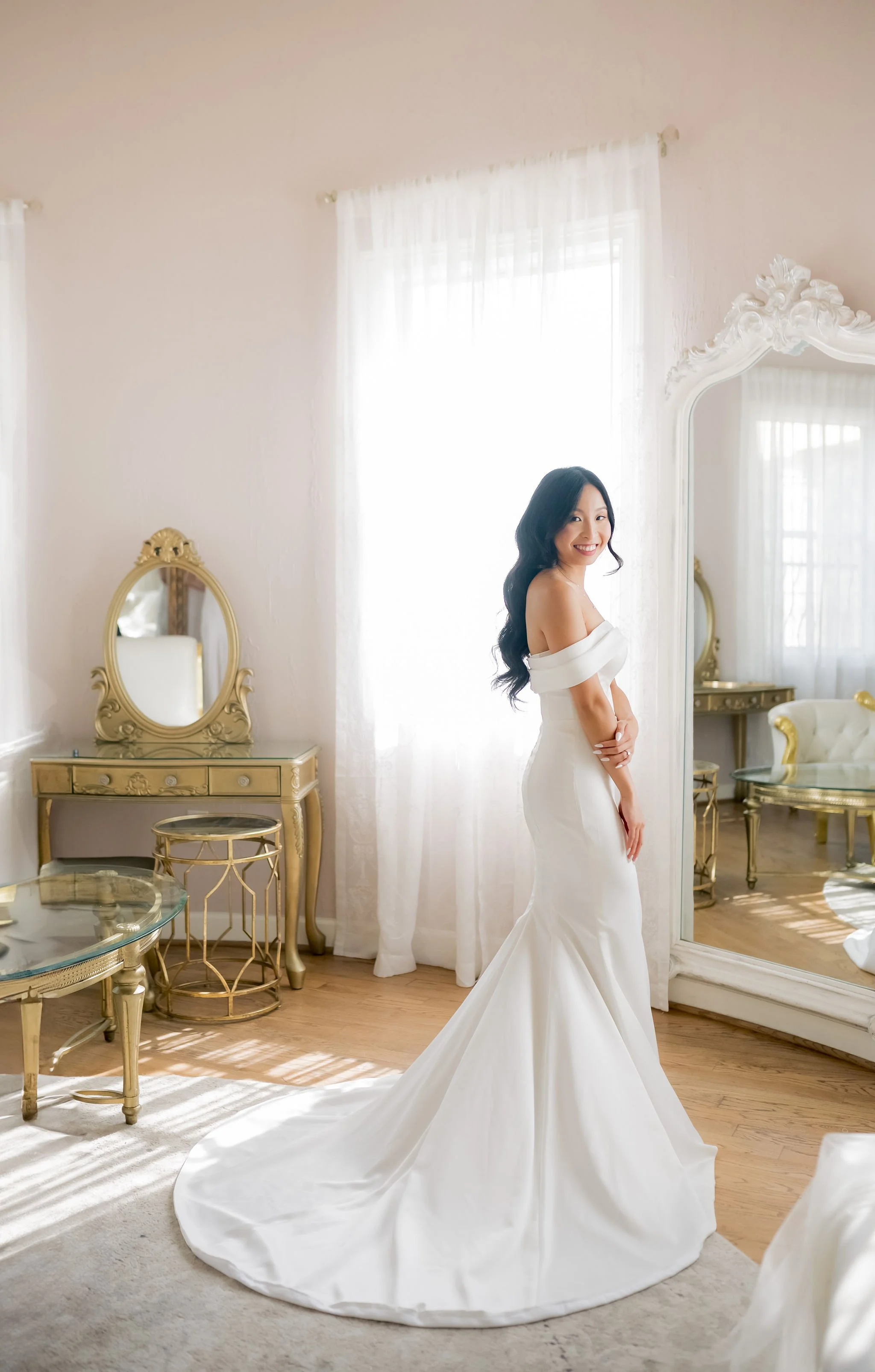 A woman in a white wedding dress standing in a bright, elegant room with gold and mirror decor, smiling.