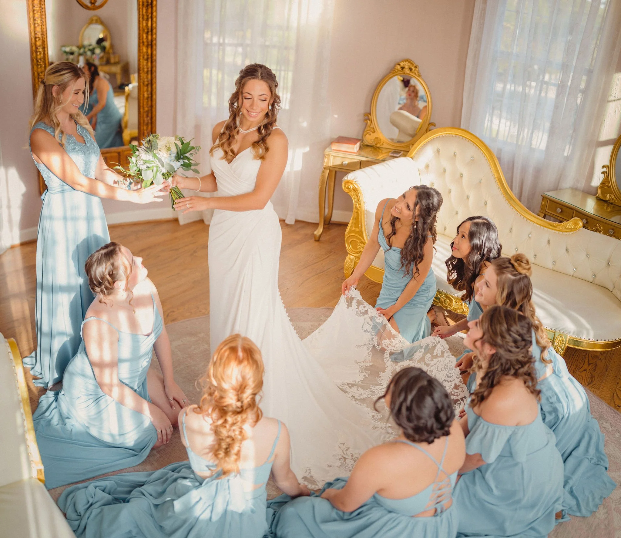 Bride receiving a bouquet of flowers from a bridesmaid, surrounded by bridesmaids in blue dresses, in a decorated room with gold furniture.