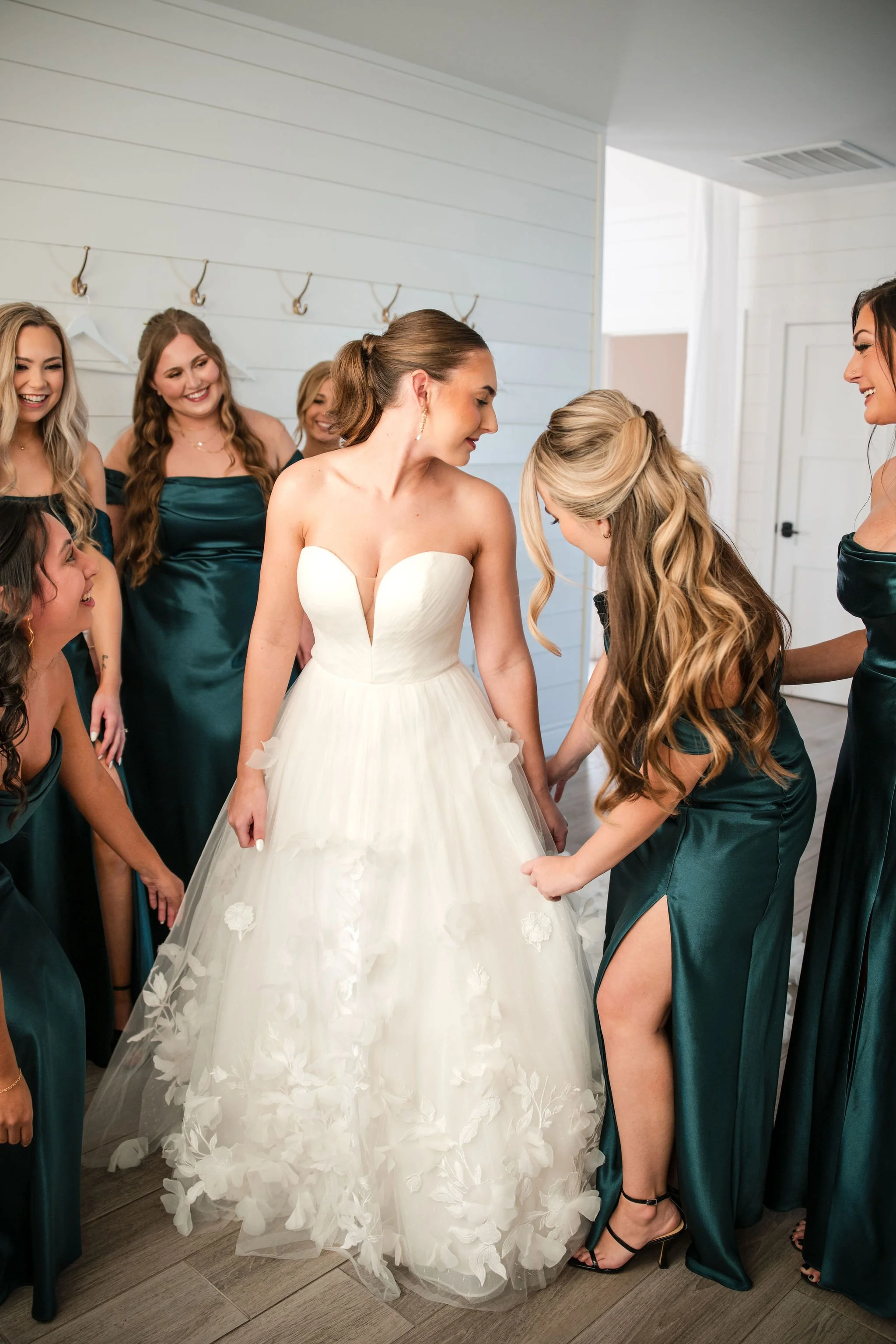 Bride in a white wedding dress surrounded by bridesmaids in teal dresses during dress fitting.