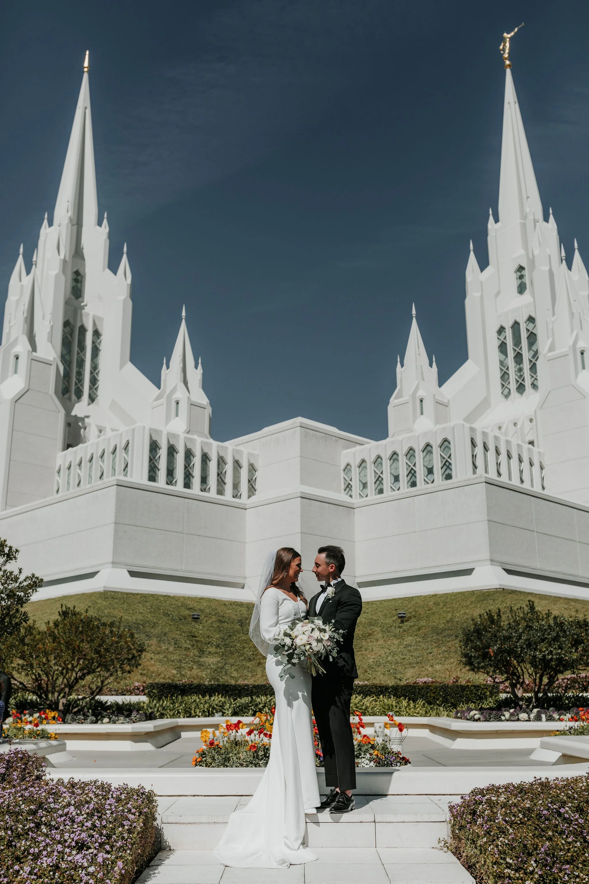 A bride and groom stand close together, smiling and holding a bouquet of flowers, in front of a large white church with tall steeples.