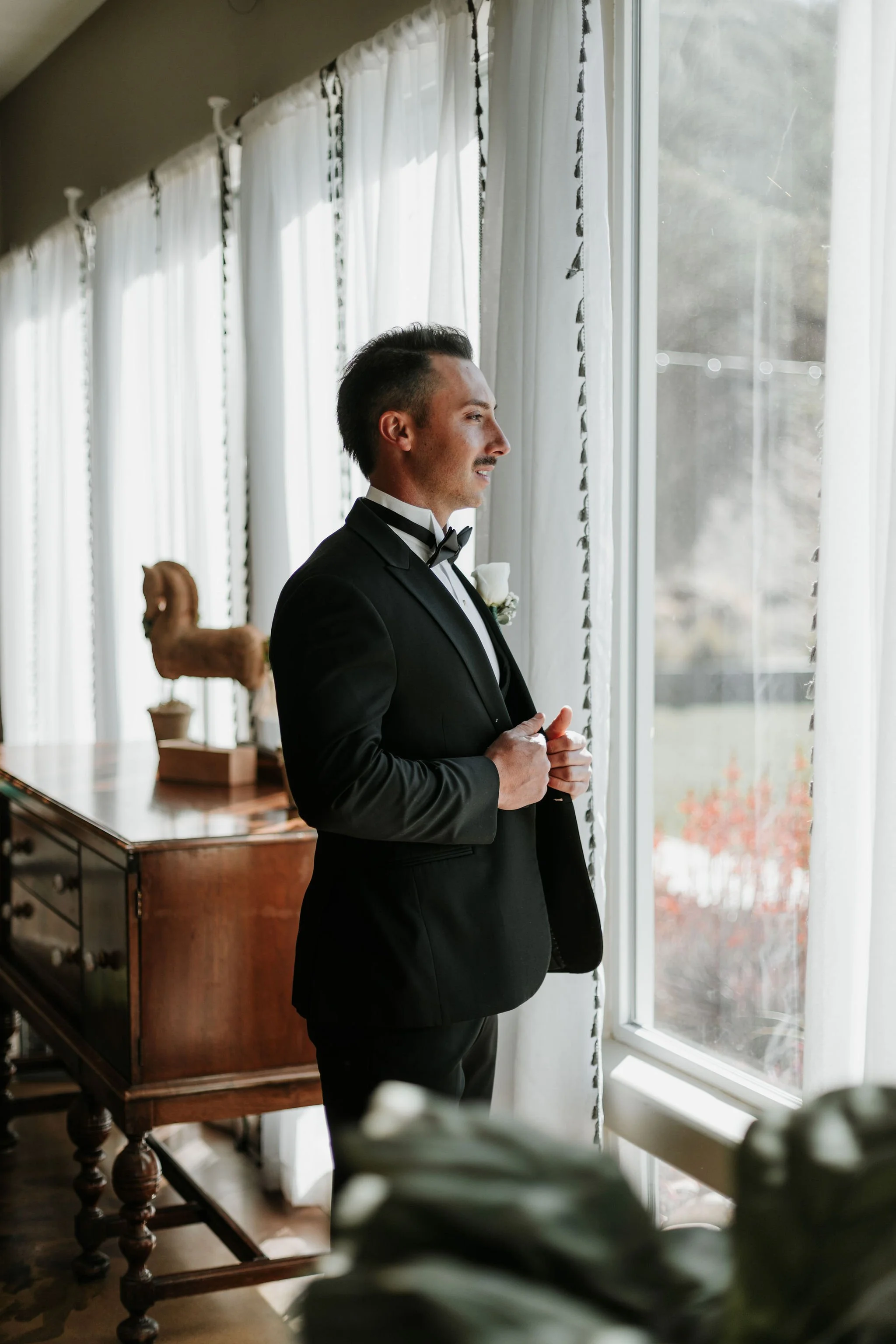 A man in a black tuxedo with a bowtie, standing by a window, looking outside.