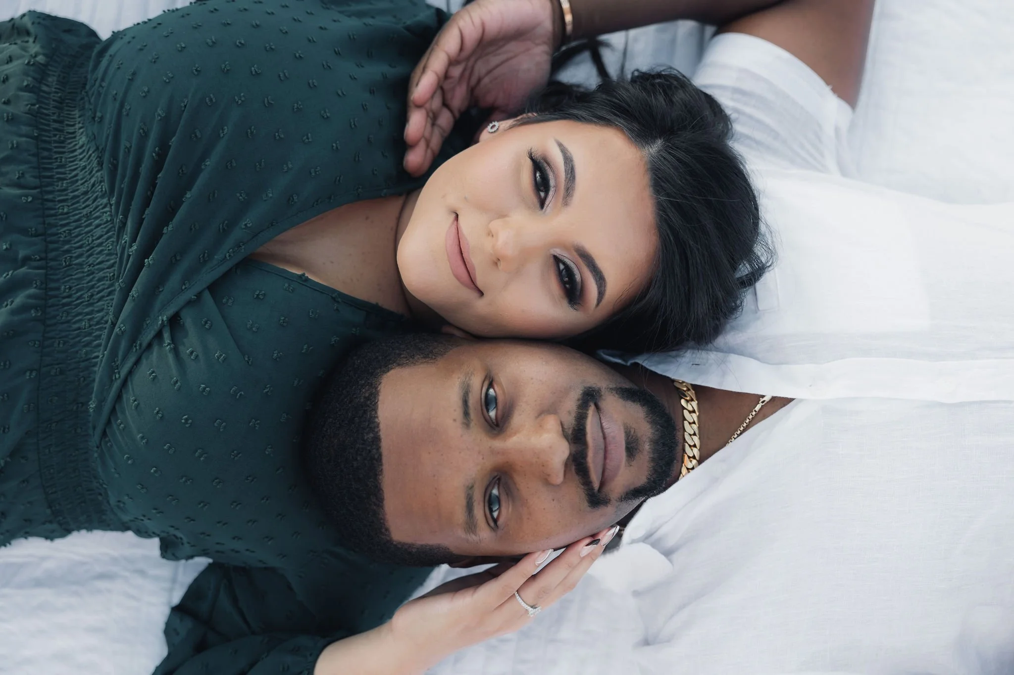 A smiling woman with dark hair and makeup lying on a bed with a man with a beard, wearing a white shirt and gold chain, both resting their heads together on a white pillow.