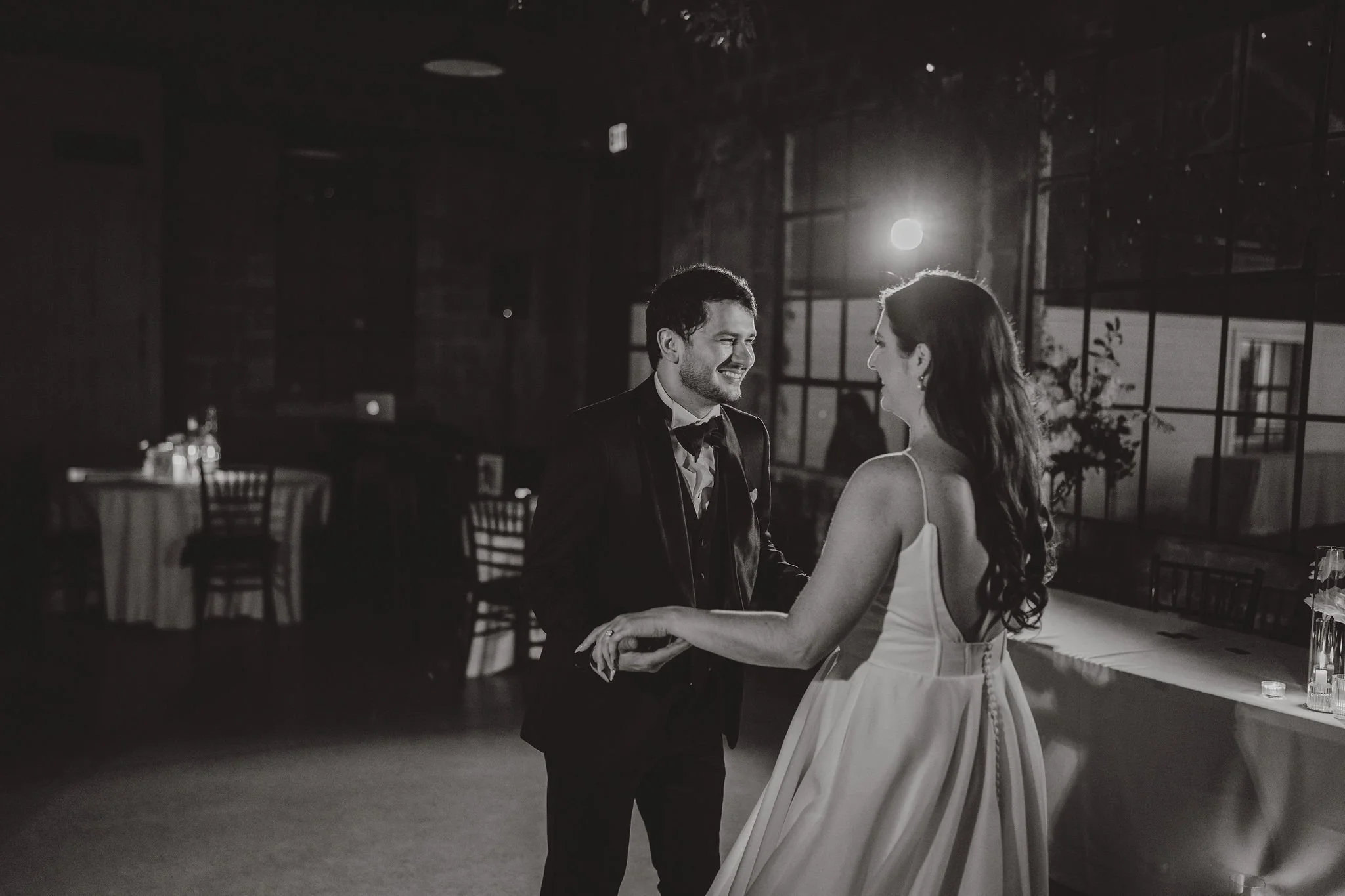 Black and white photo of a bride and groom dancing together at their wedding reception, holding hands and smiling at each other, with tables and chairs in the background.