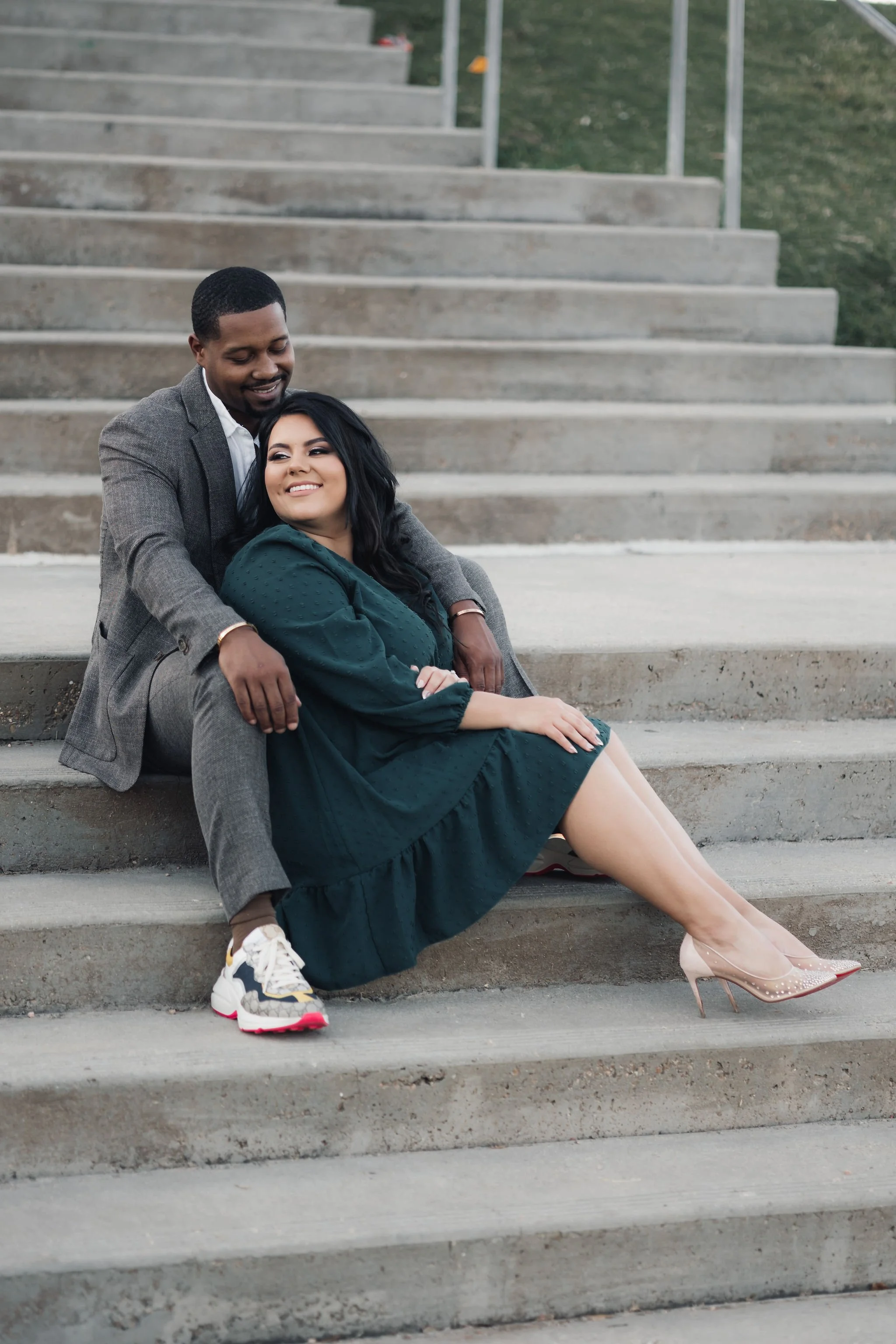 A man and a woman sitting together on concrete steps outdoors, smiling. The woman is wearing a dark green dress and high heels, while the man is dressed in a gray suit and sneakers.