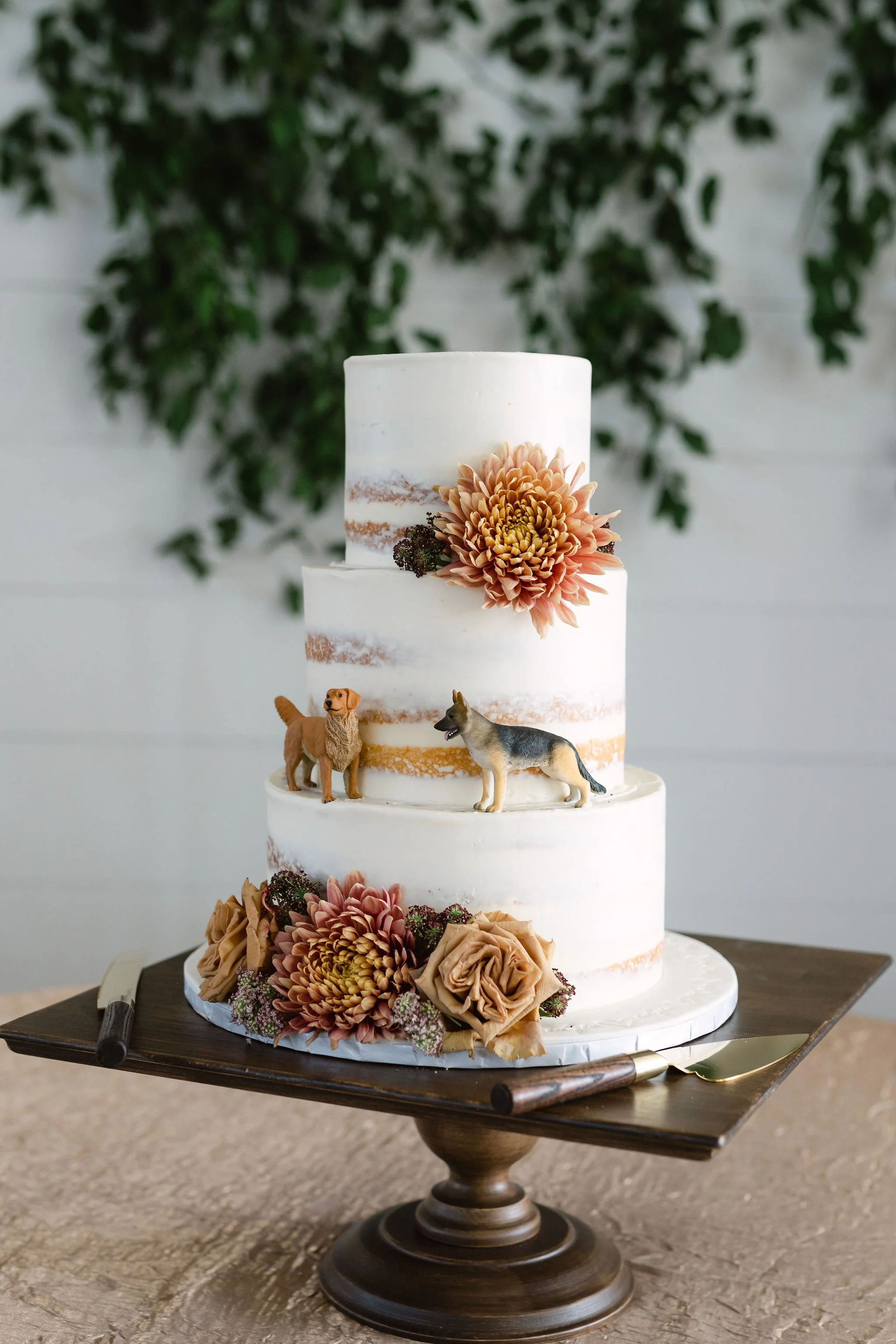 Three-tiered wedding cake with semi-naked frosting, decorated with pink and beige flowers, and dog figurines on the middle tier. The cake is on a wooden cake stand with a cake knife and server beside it.