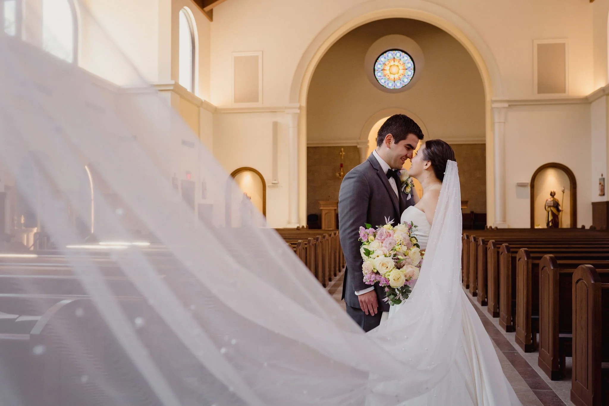 A bride and groom share a moment in a church, with the bride holding a bouquet of flowers and wearing a white wedding gown with a veil, and the groom in a dark tuxedo.